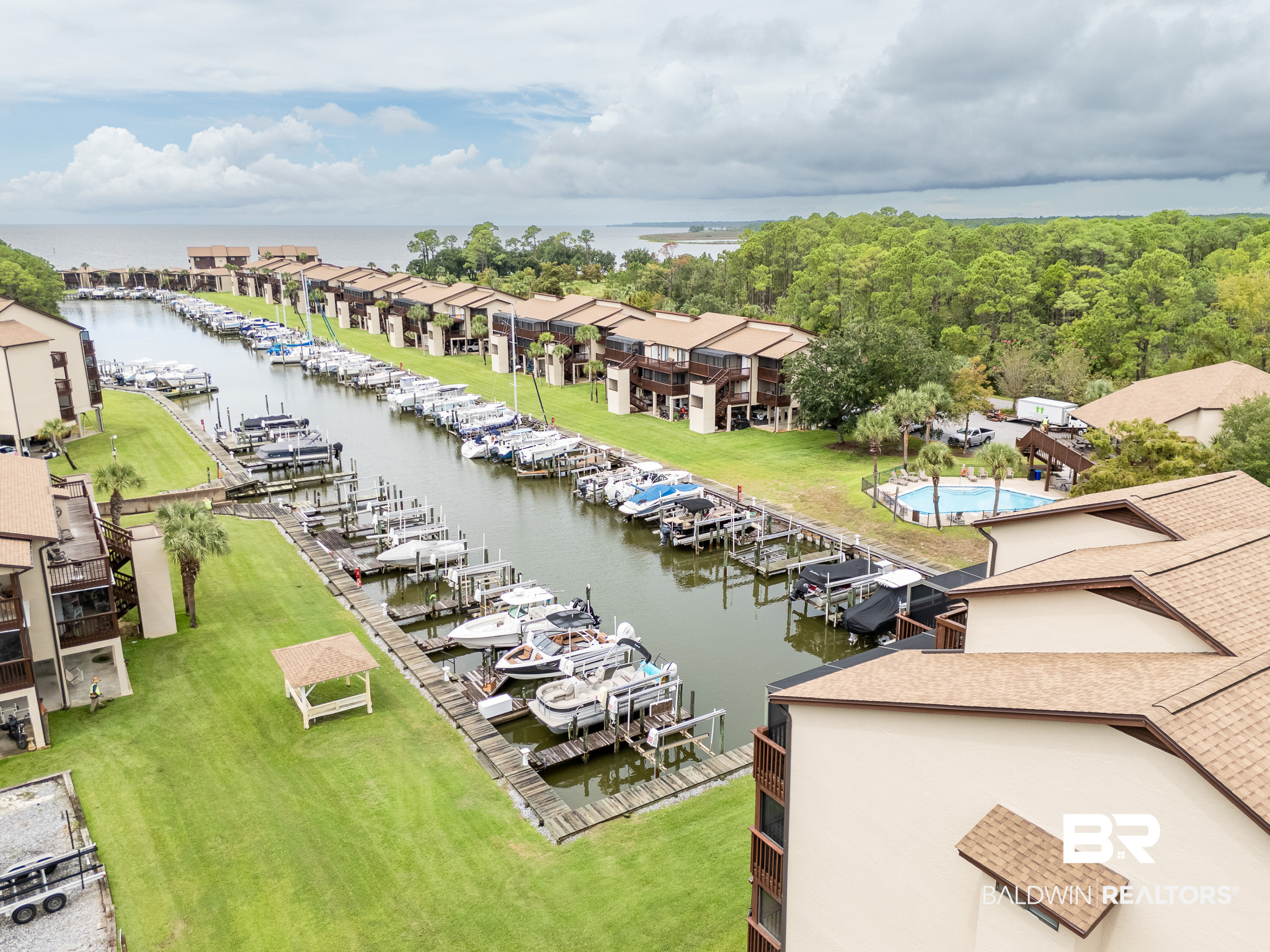 Aerial view of waterfront condominiums with a marina, docked boats, swimming pool, and wooded shoreline.