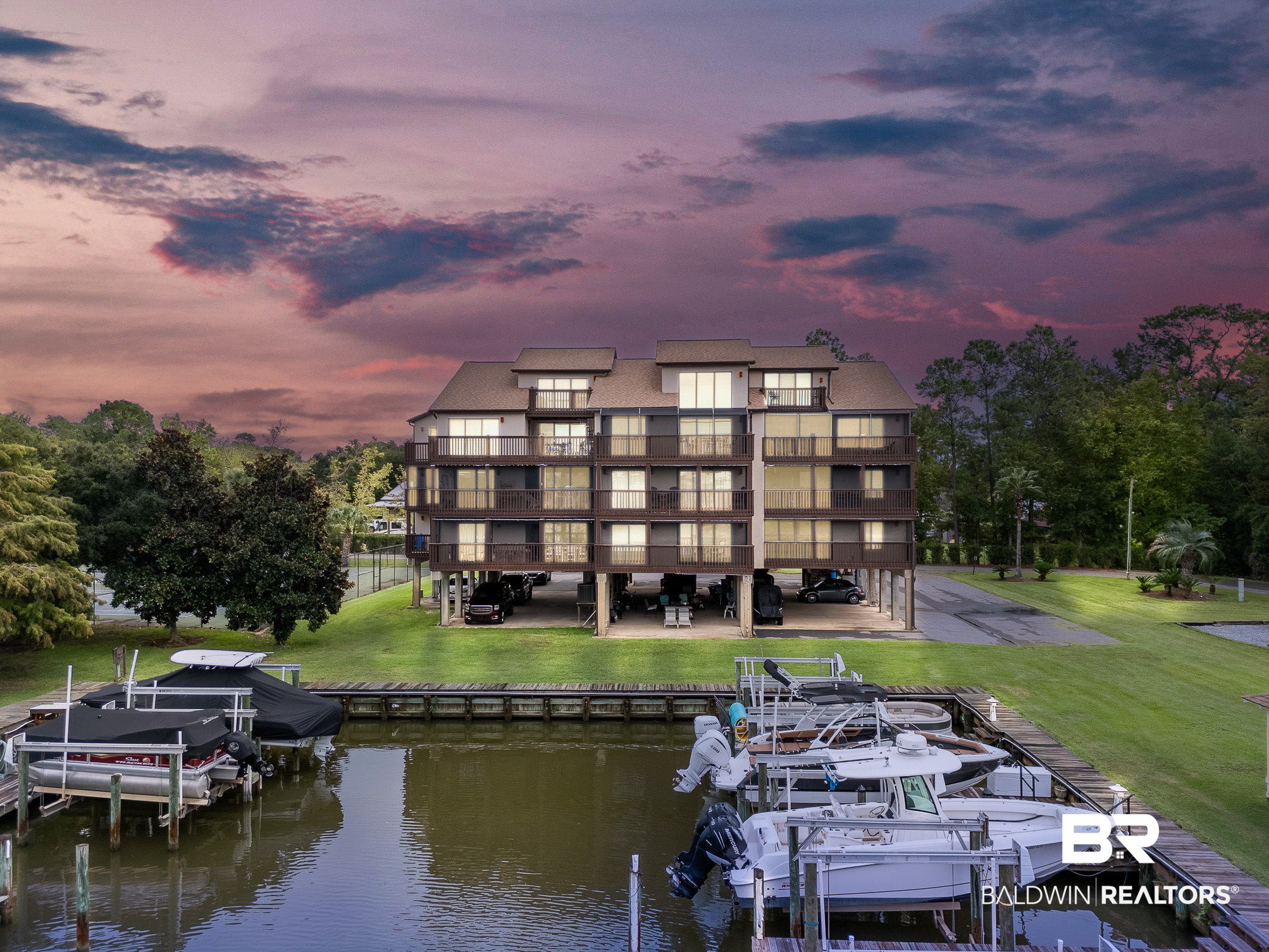 Waterfront condominium with tiered balconies and boats docked in front beneath a pink dusk sky.