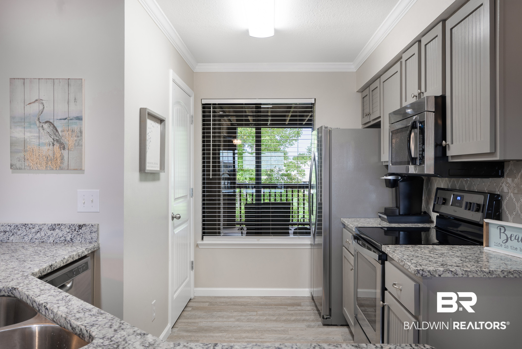 Condominium kitchen with gray cabinetry, granite counters, stainless steel appliances, and a window overlooking greenery.