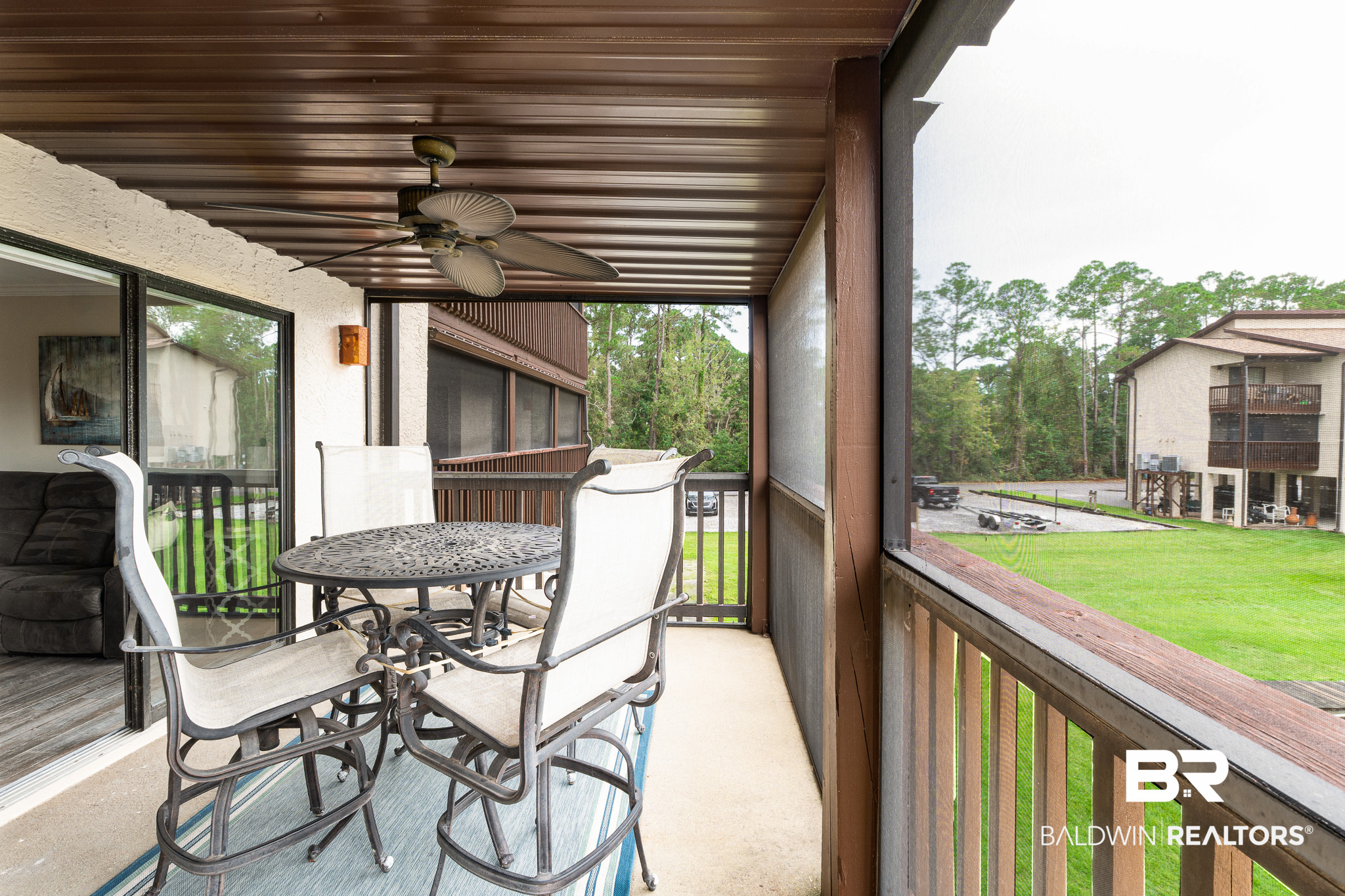 Covered balcony with outdoor dining set and ceiling fan overlooking a green courtyard and neighboring building.