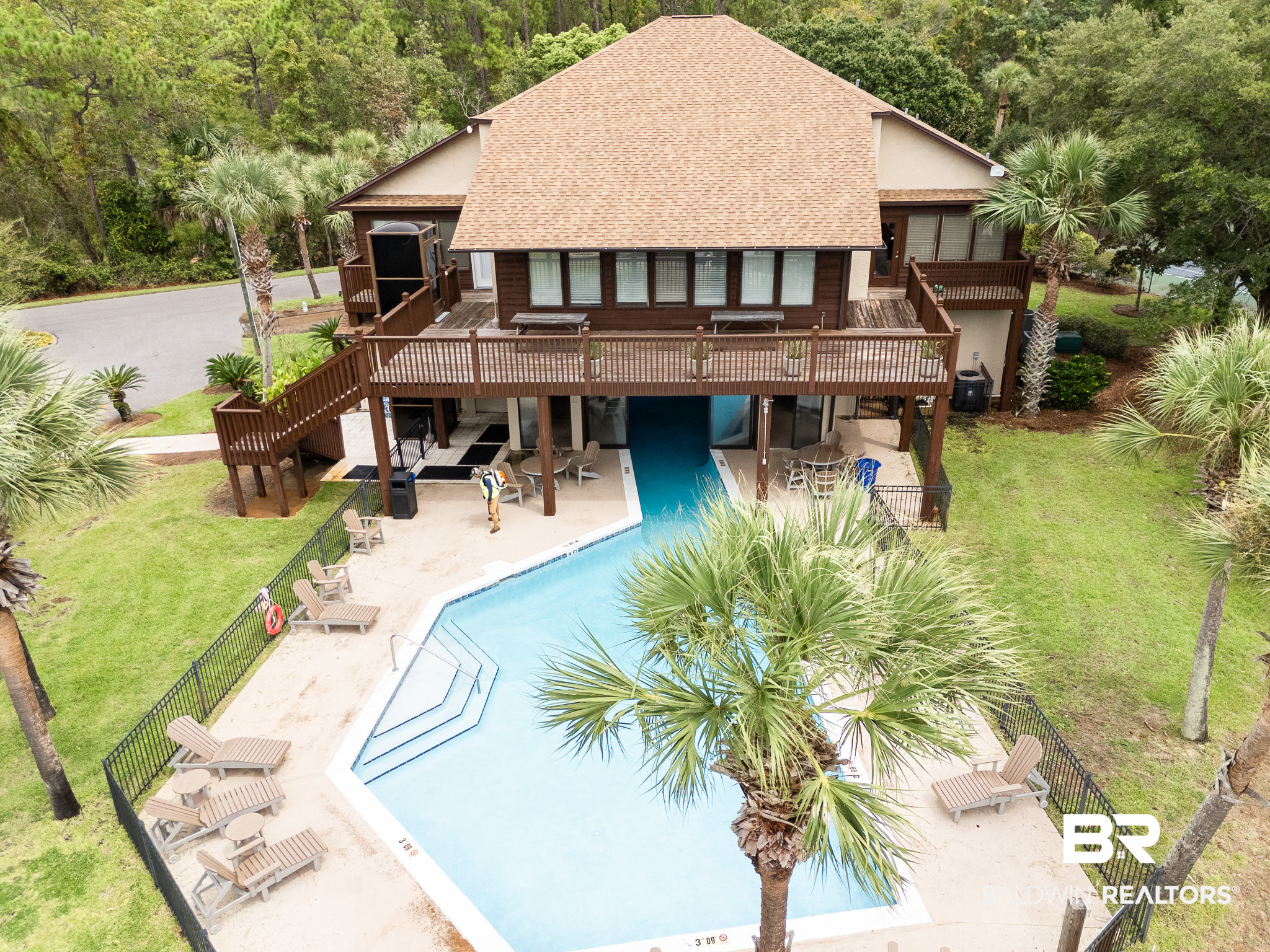 Elevated view of a home with expansive wooden decks overlooking a fenced swimming pool and palm trees.