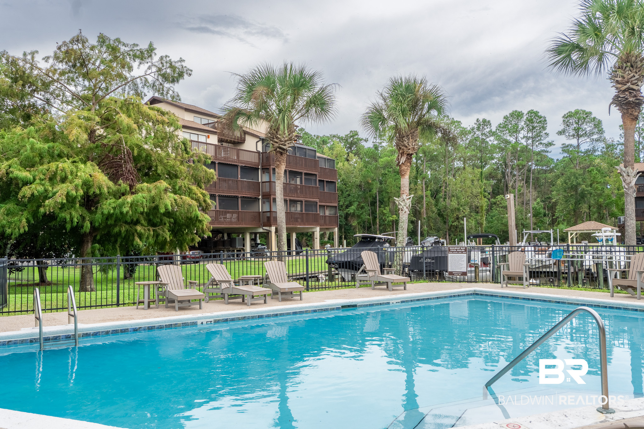 Outdoor condominium pool with lounge chairs, palm trees, and multi-story building in the background.