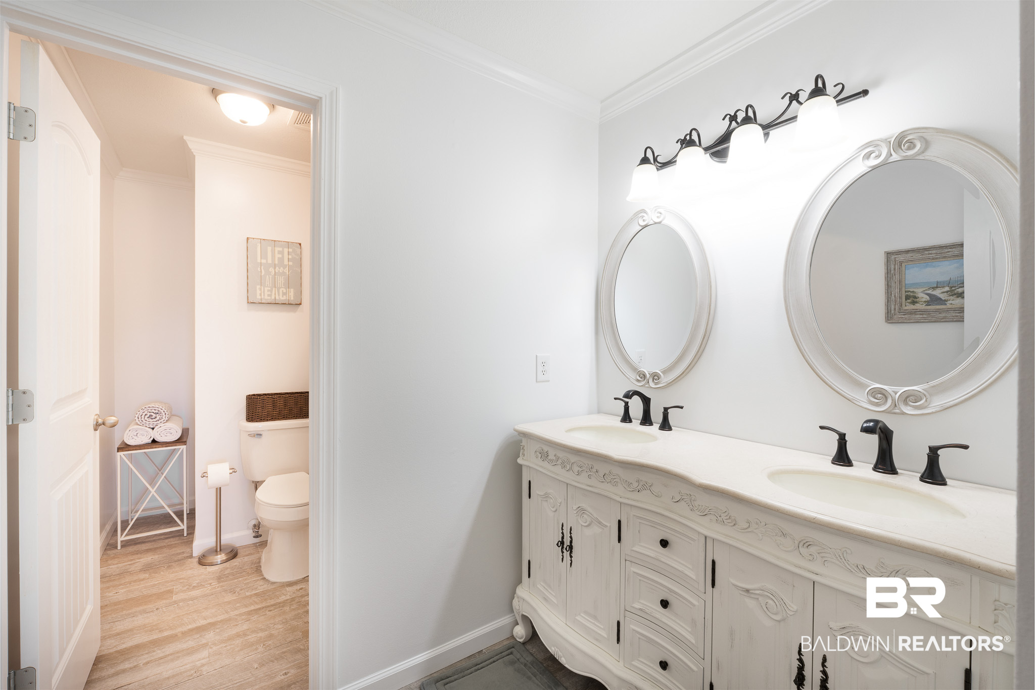 Bathroom with an ornate double vanity, oval mirrors, black faucets, and an adjoining toilet nook.