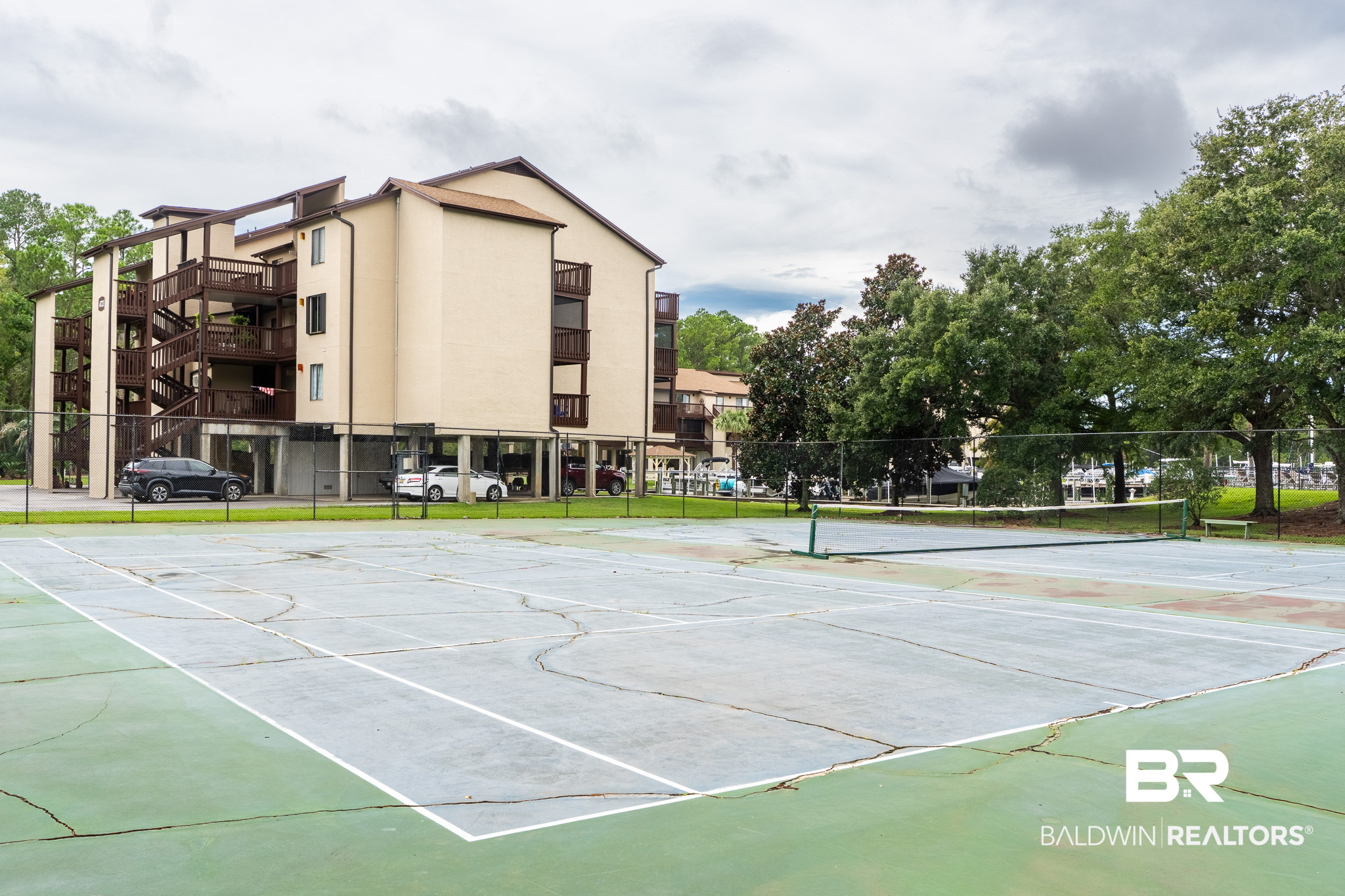 Condominium building behind a fenced tennis court, with dark wood balconies and mature trees.