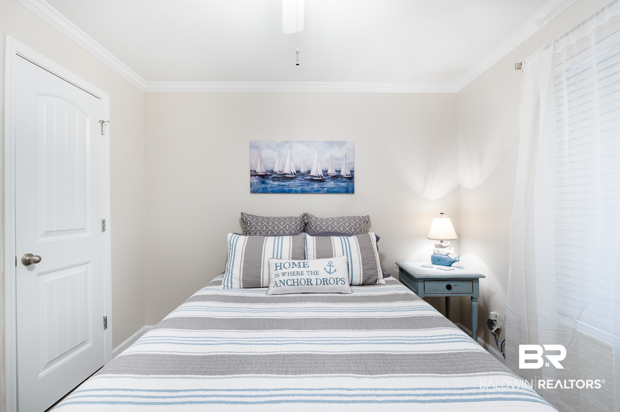 Bedroom with striped bedding, a sailboat painting, and a cushion reading “Home is where the anchor drops.”