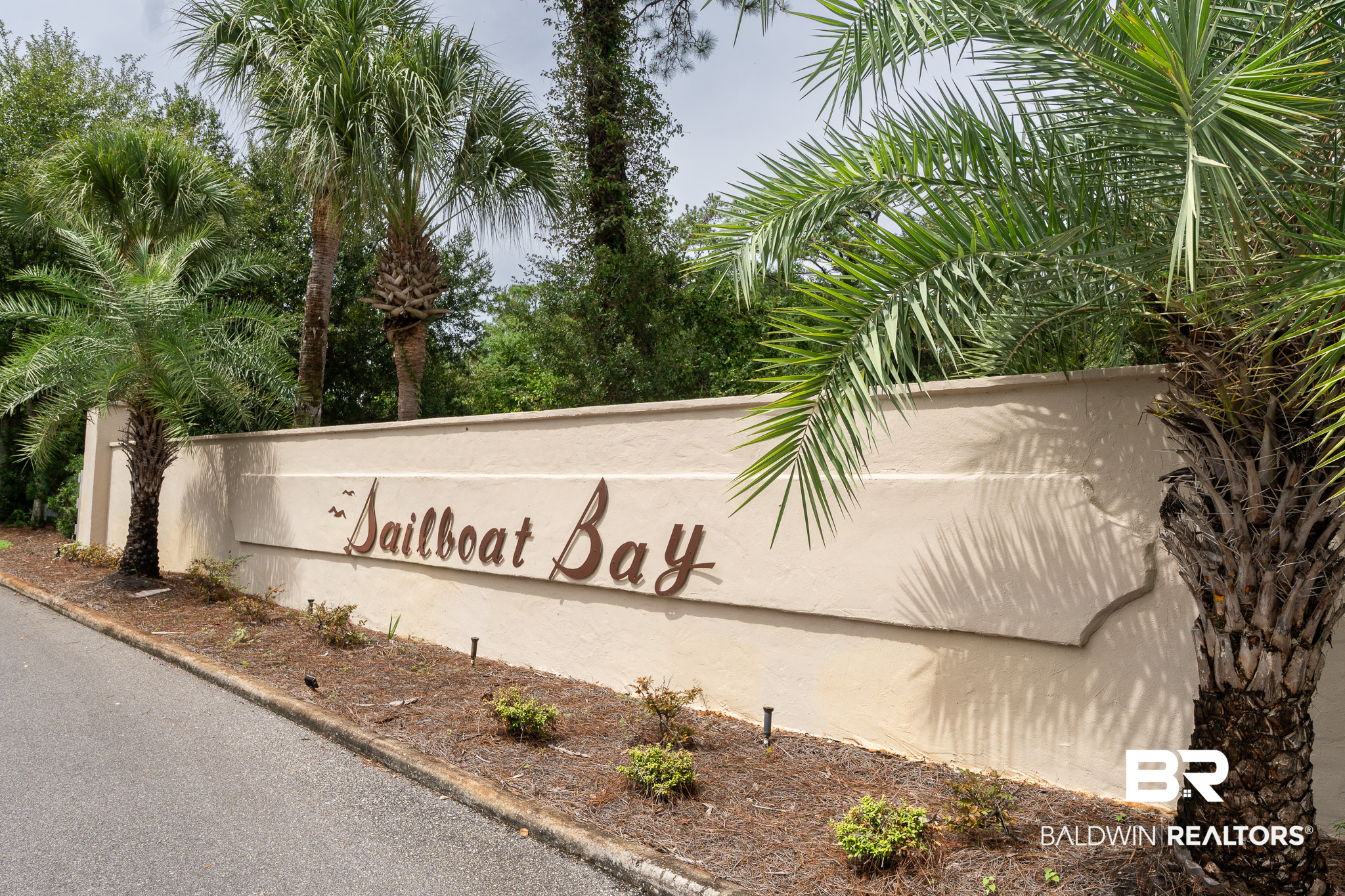 Sailboat Bay entrance sign on a stucco wall framed by palm trees.