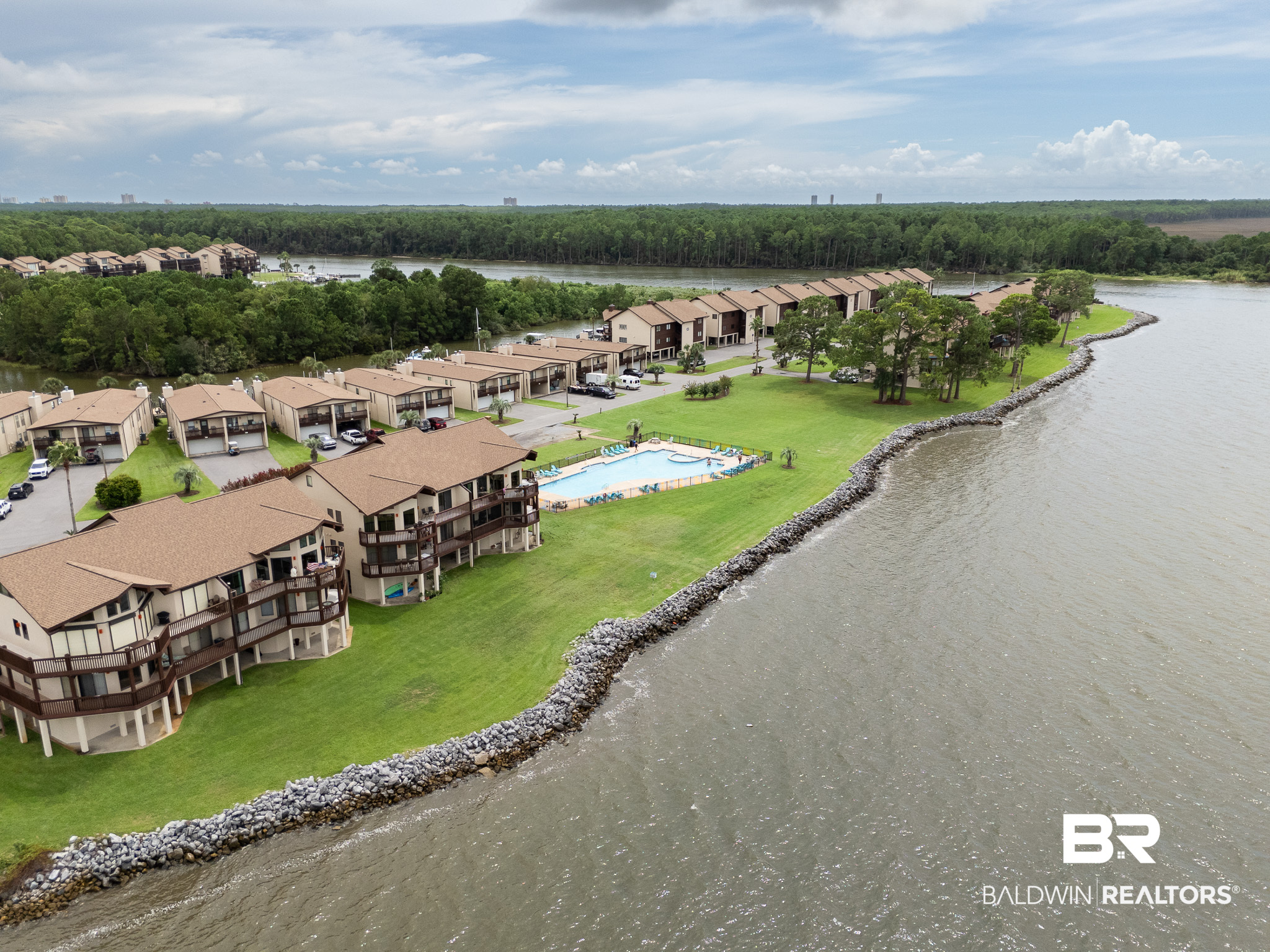 Waterfront condominium community with a pool, green lawn, and rocky shoreline.