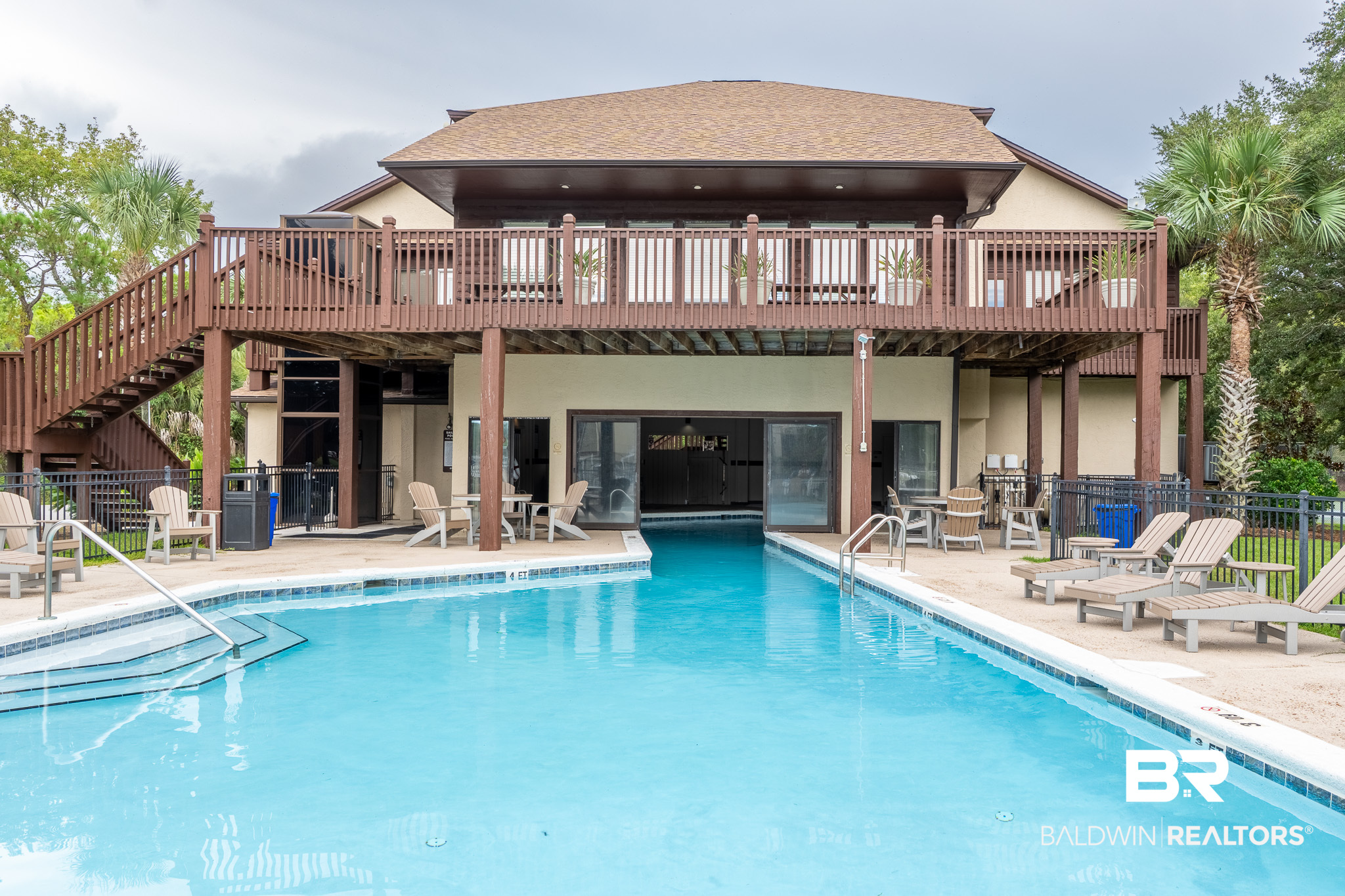 Outdoor pool in front of a raised building with a wide wooden deck and surrounding lounge chairs.