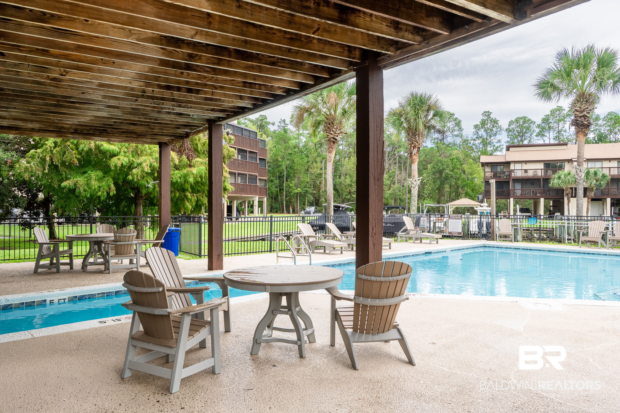 Community pool beneath a wooden pergola, with Adirondack chairs and palm trees.