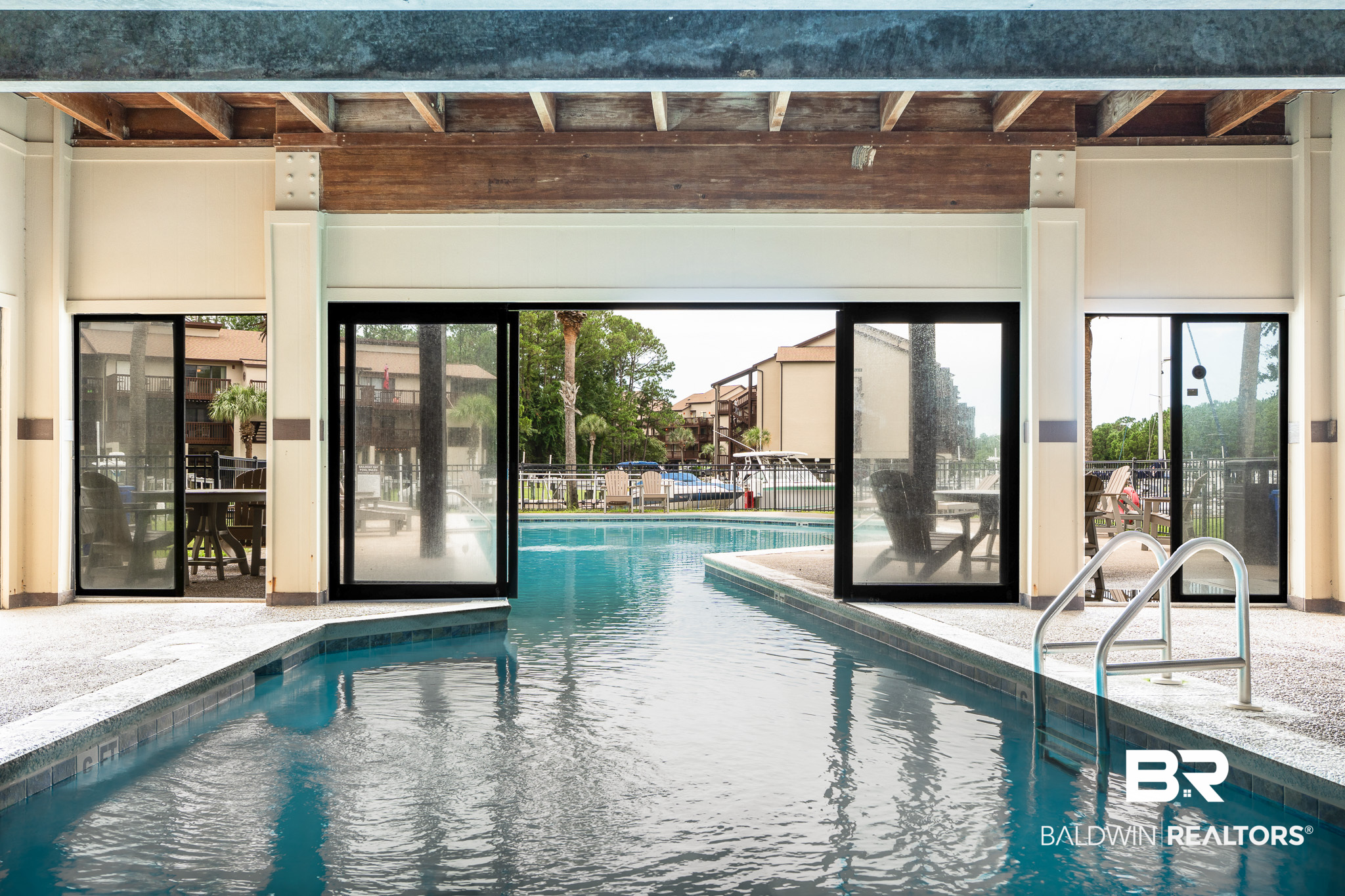 Covered swimming pool with patio seating and neighboring condominium buildings beyond the glass doors.