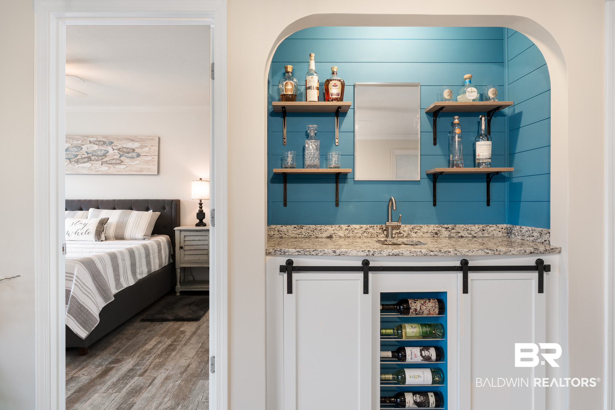 Blue wet bar with granite counter, open bottle shelves, and built-in wine rack beside a bedroom doorway.
