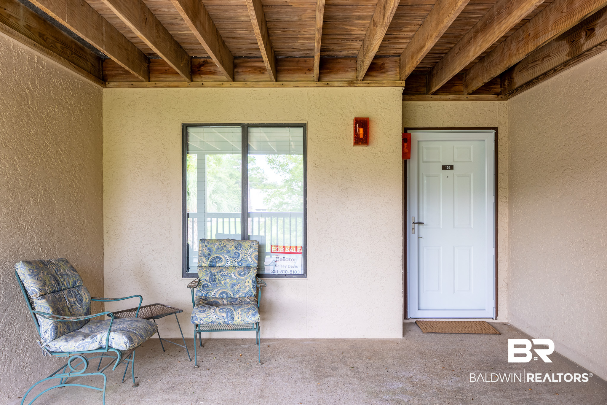 Covered porch with exposed timber ceiling, two patterned chairs, and the entrance to Unit 102.