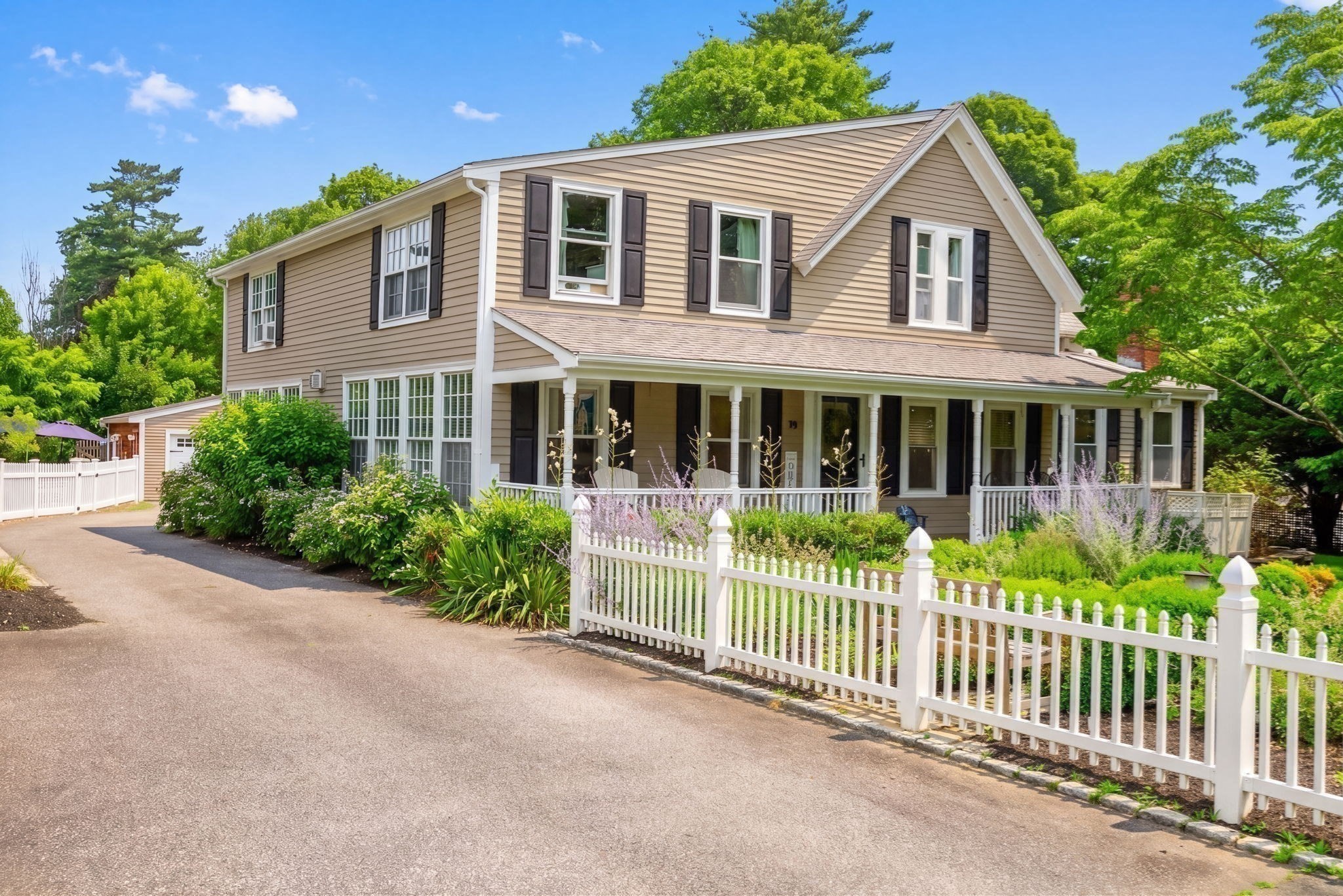Two-story beige home with a long covered porch, white picket fence, and lush gardens.