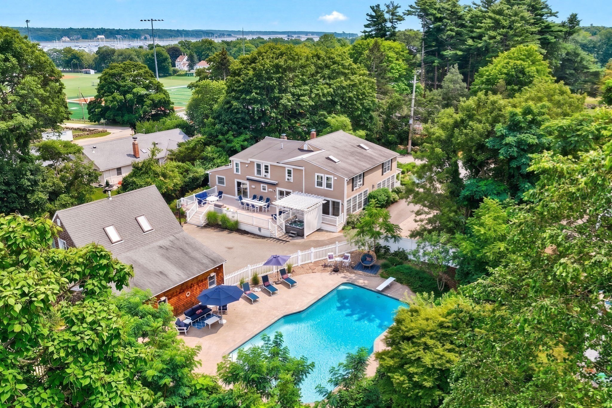 Aerial view of a tree-surrounded house with a swimming pool and distant marina.