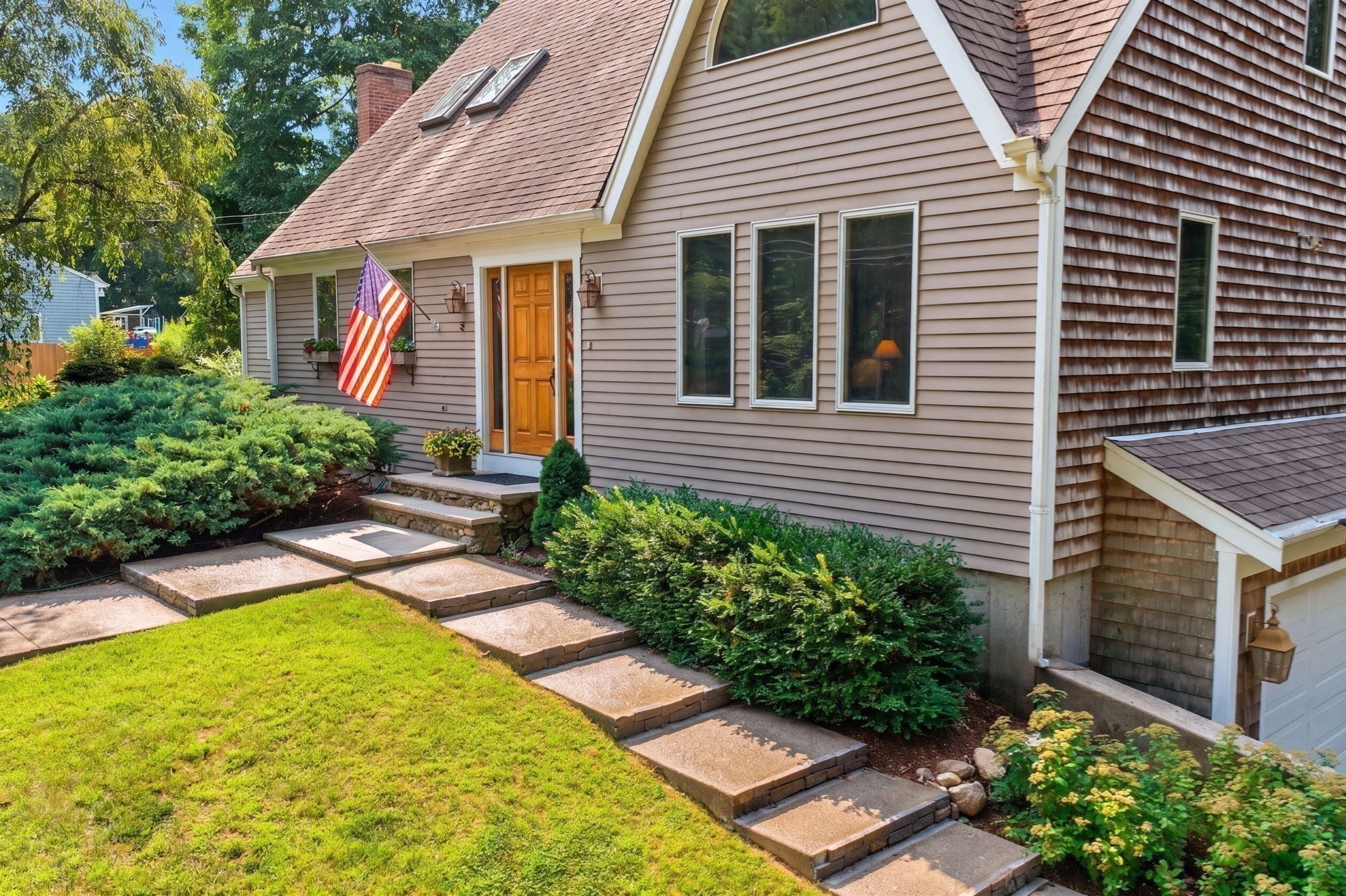 Home entrance with a warm wood door, stone-edged steps, landscaped shrubs, and an American flag.