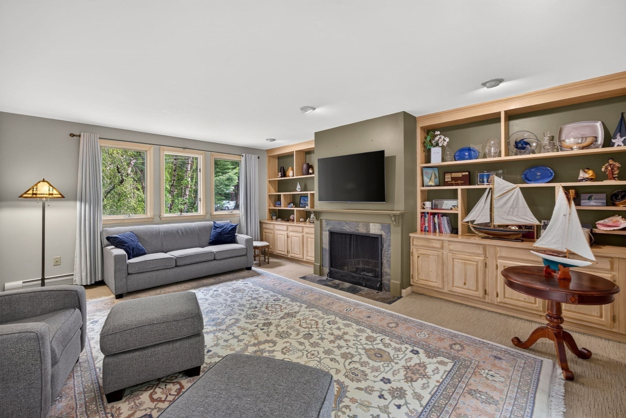 Living room with a fireplace, built-in display shelves, gray seating, and windows overlooking trees.