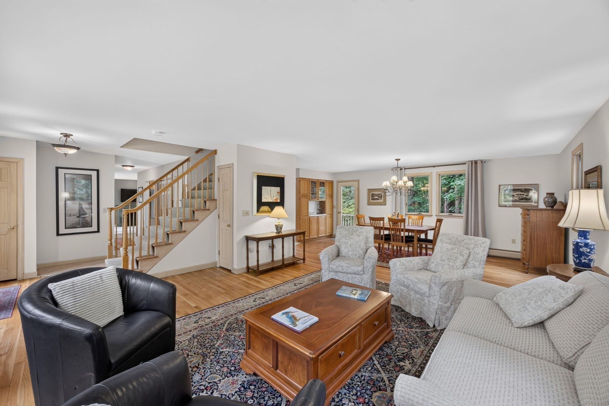 Furnished living room and dining area with a broad staircase, hardwood floors, and large windows.
