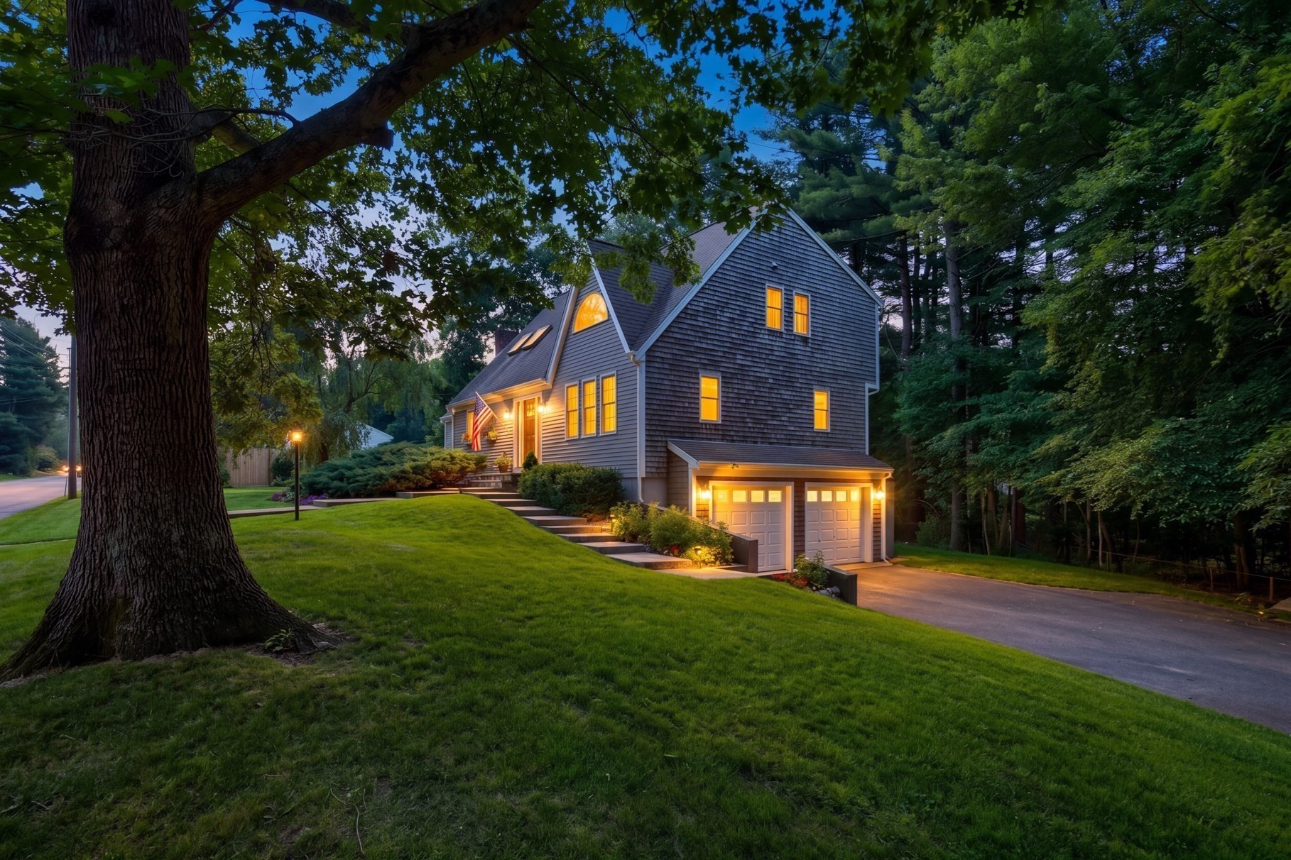 Gray-shingle home with warmly lit windows and a two-car garage, framed by a large tree and broad lawn at dusk.