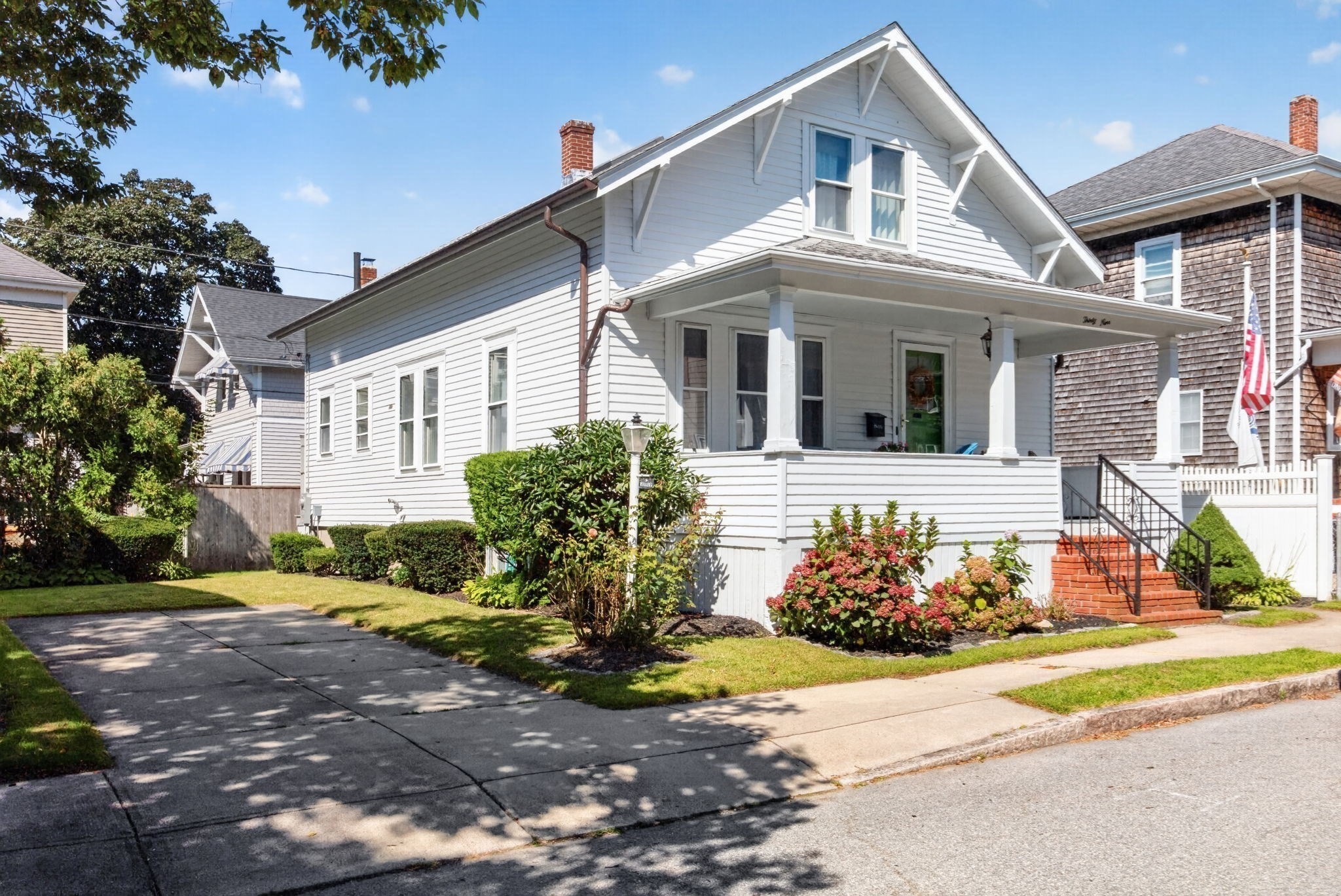 White gable-front house with a covered porch, brick steps, and flowering shrubs beside the driveway.