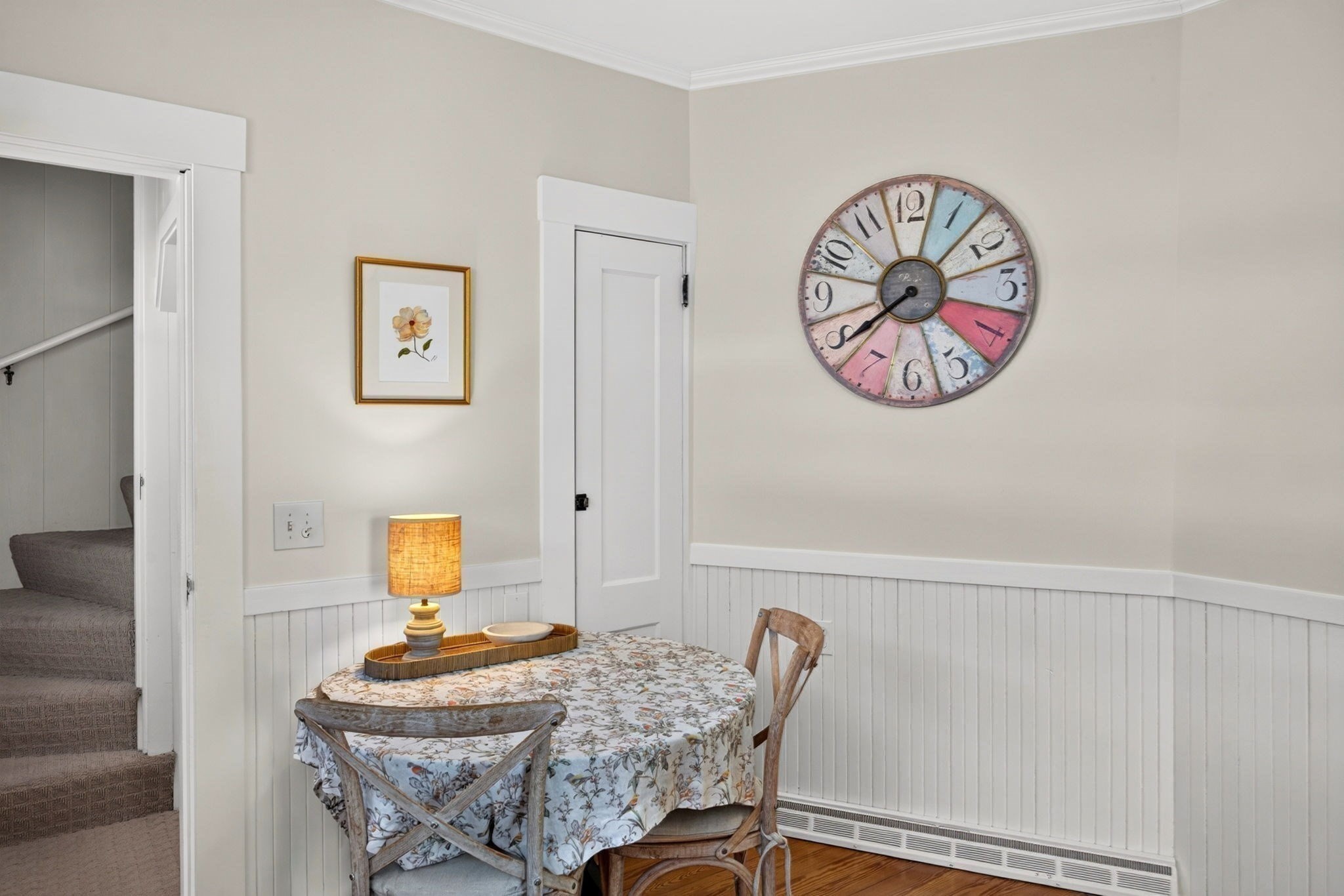 Dining area with a floral-covered round table, beadboard wainscoting, and a large colorful wall clock.