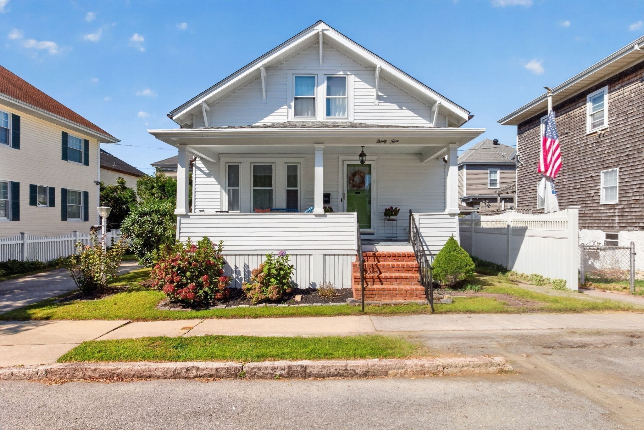 White two-story house with a covered porch, green door, brick steps, and flowering shrubs.