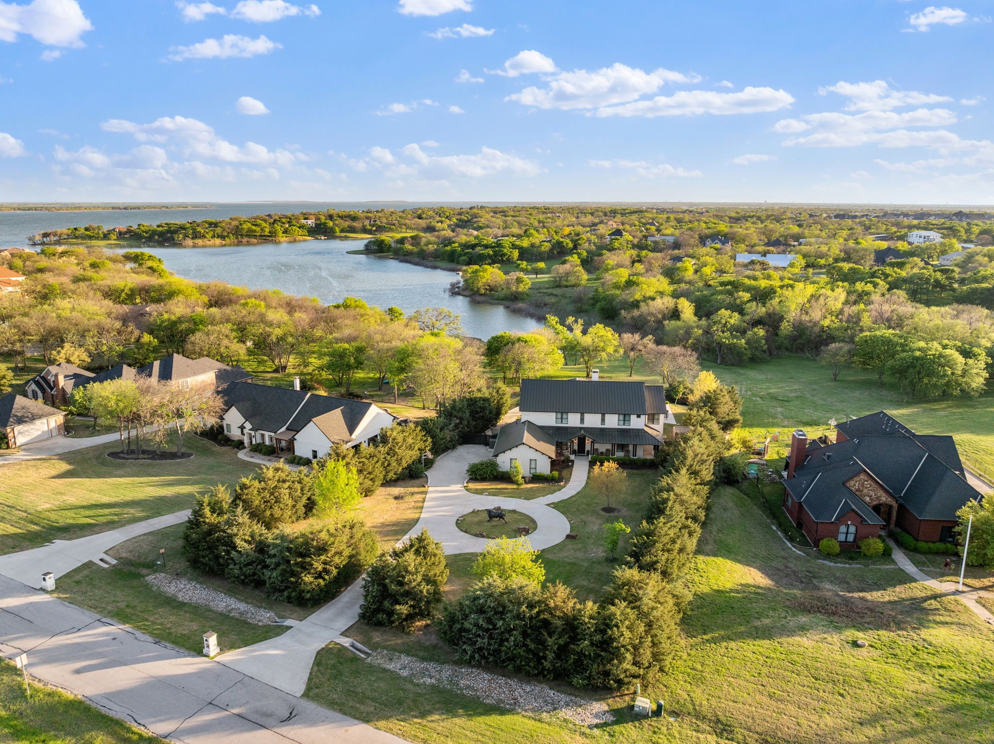 Aerial view of a two-story home beside a lake, with a curved driveway and mature trees.