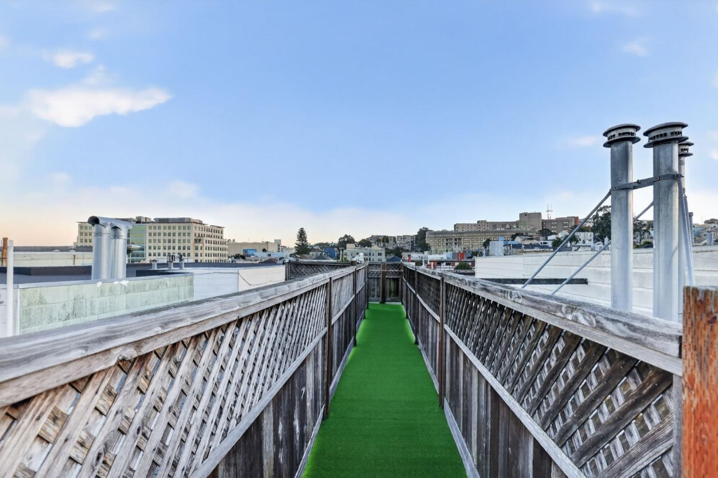 Rooftop deck with a green-turf walkway, weathered wood railings, and distant city buildings.