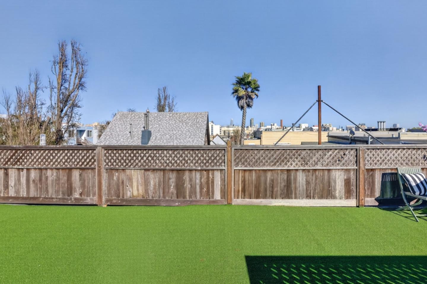 Backyard with artificial turf, a wooden fence, and a lounge chair, with a palm tree beyond the fence.