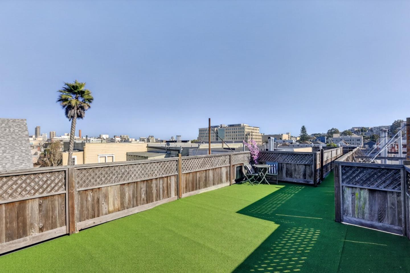Rooftop deck with green turf, wooden fencing, and a small seating area overlooking city buildings.