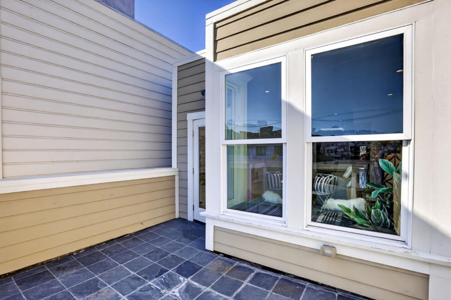 Tiled patio beside beige siding, with large windows revealing chairs and leafy plants inside.