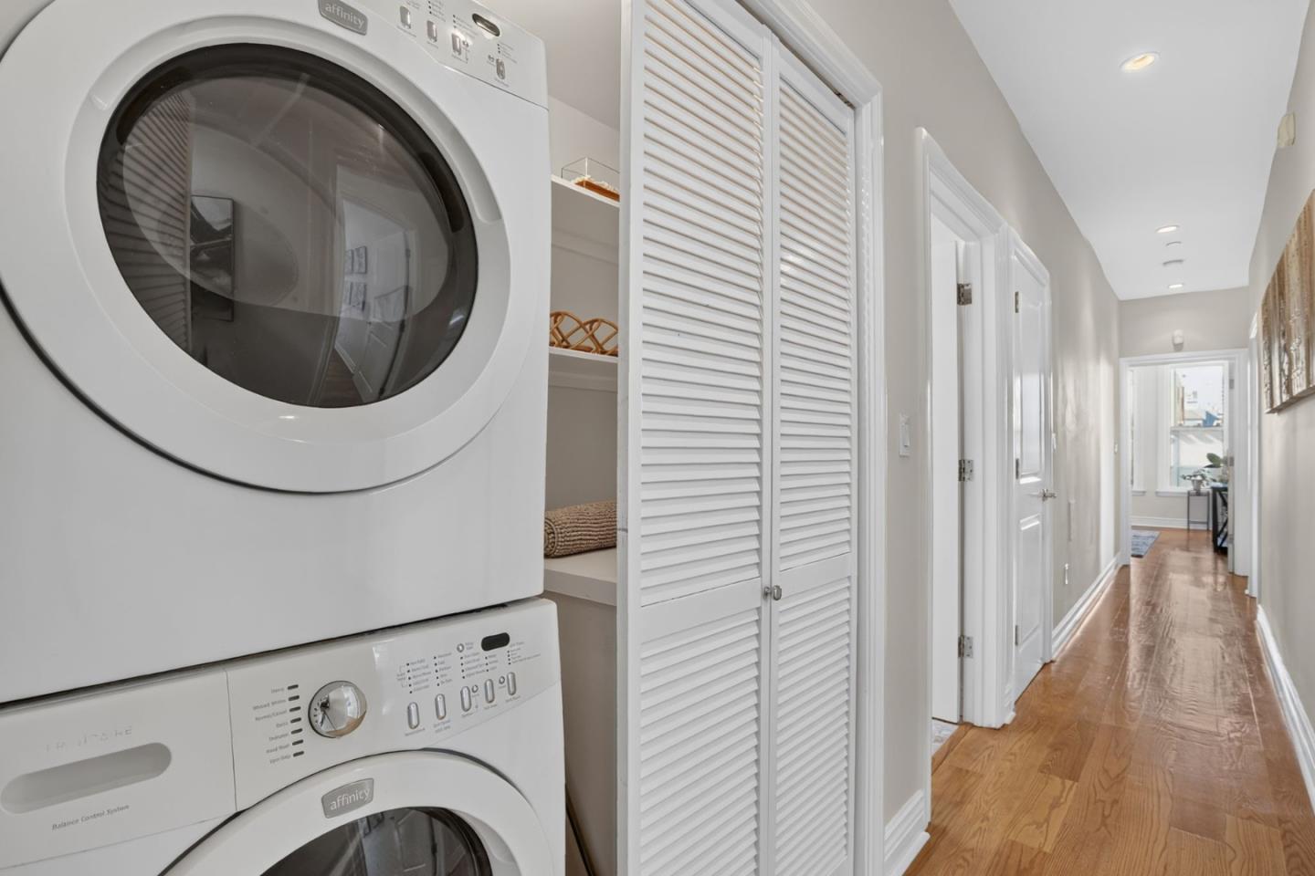 Hallway with stacked washer and dryer, louvered closet doors, and hardwood flooring.