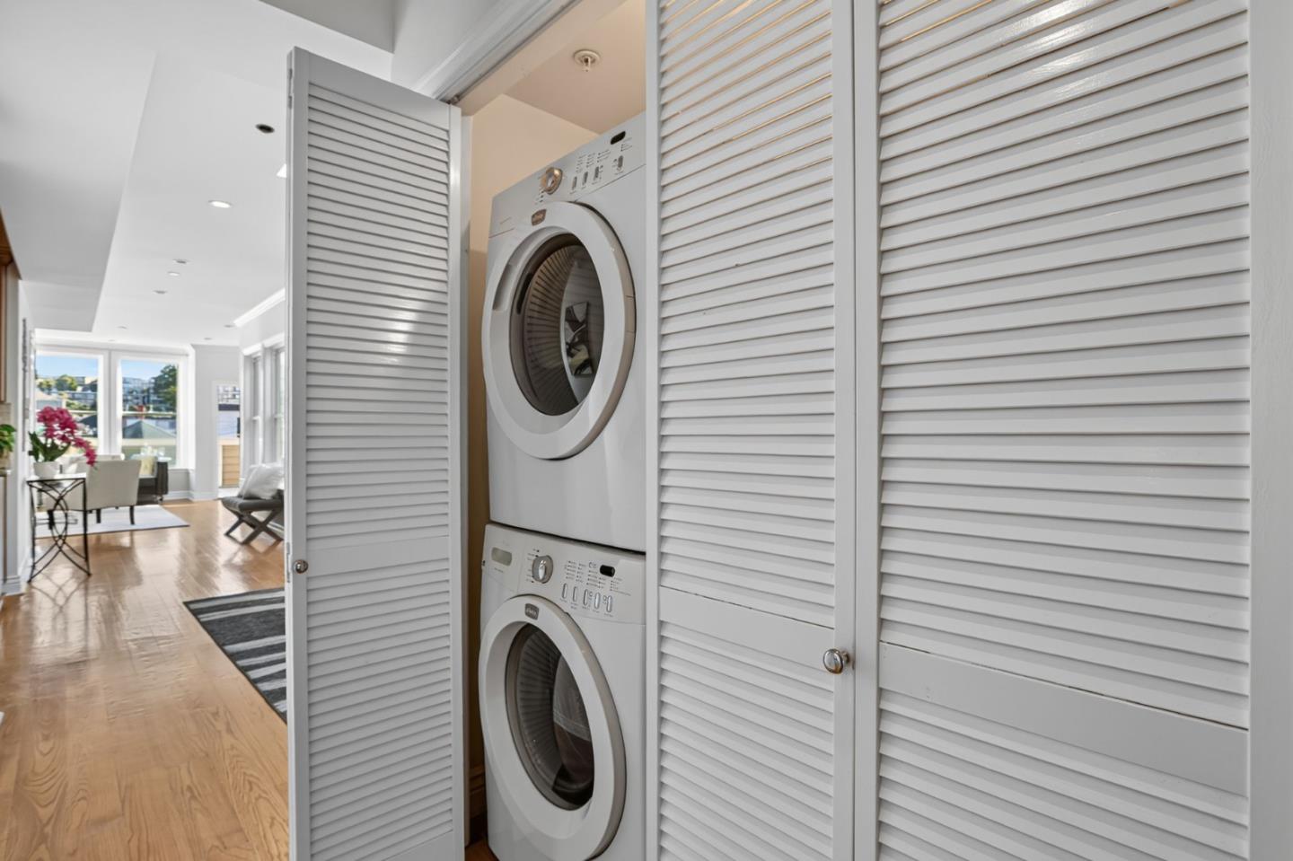 Stacked washer and dryer behind open louvered doors, with a bright living area beyond.