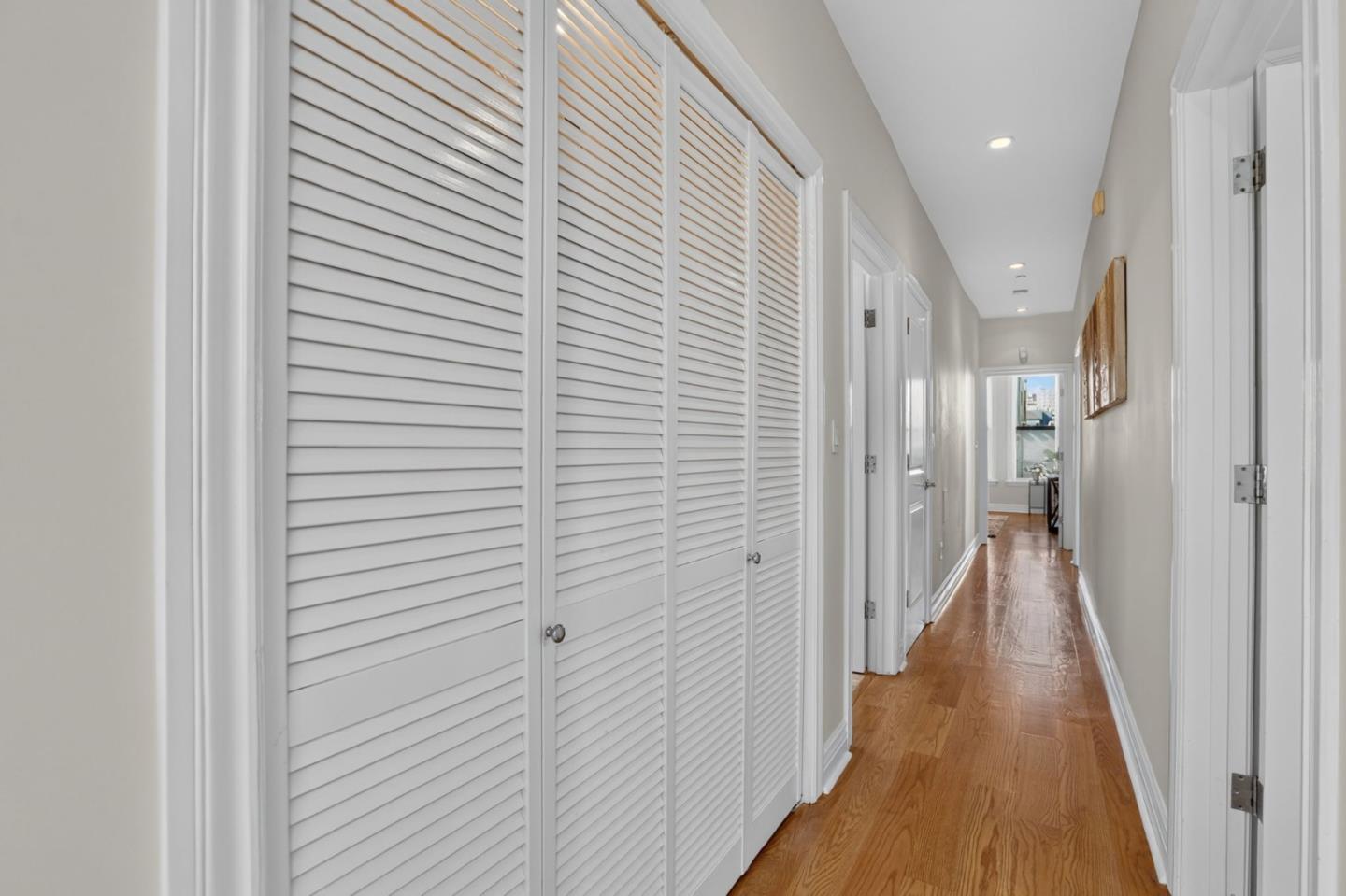 Bright hallway with hardwood floors, white louvered closet doors, and framed art.
