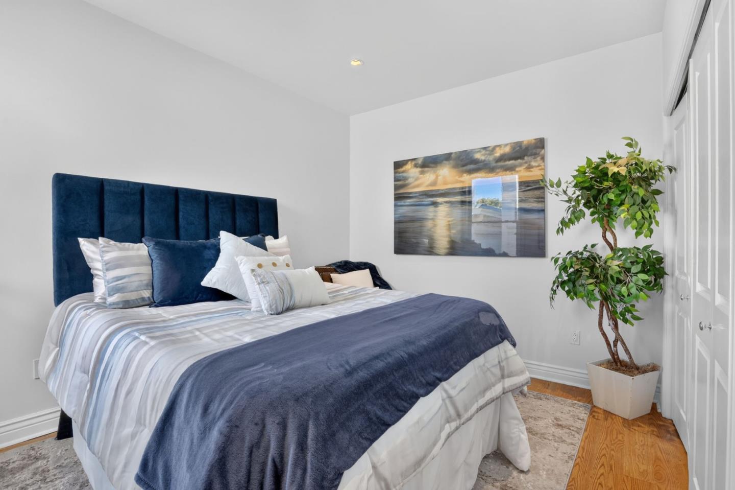 Furnished bedroom with navy headboard, striped bedding, coastal wall art, and a potted tree.