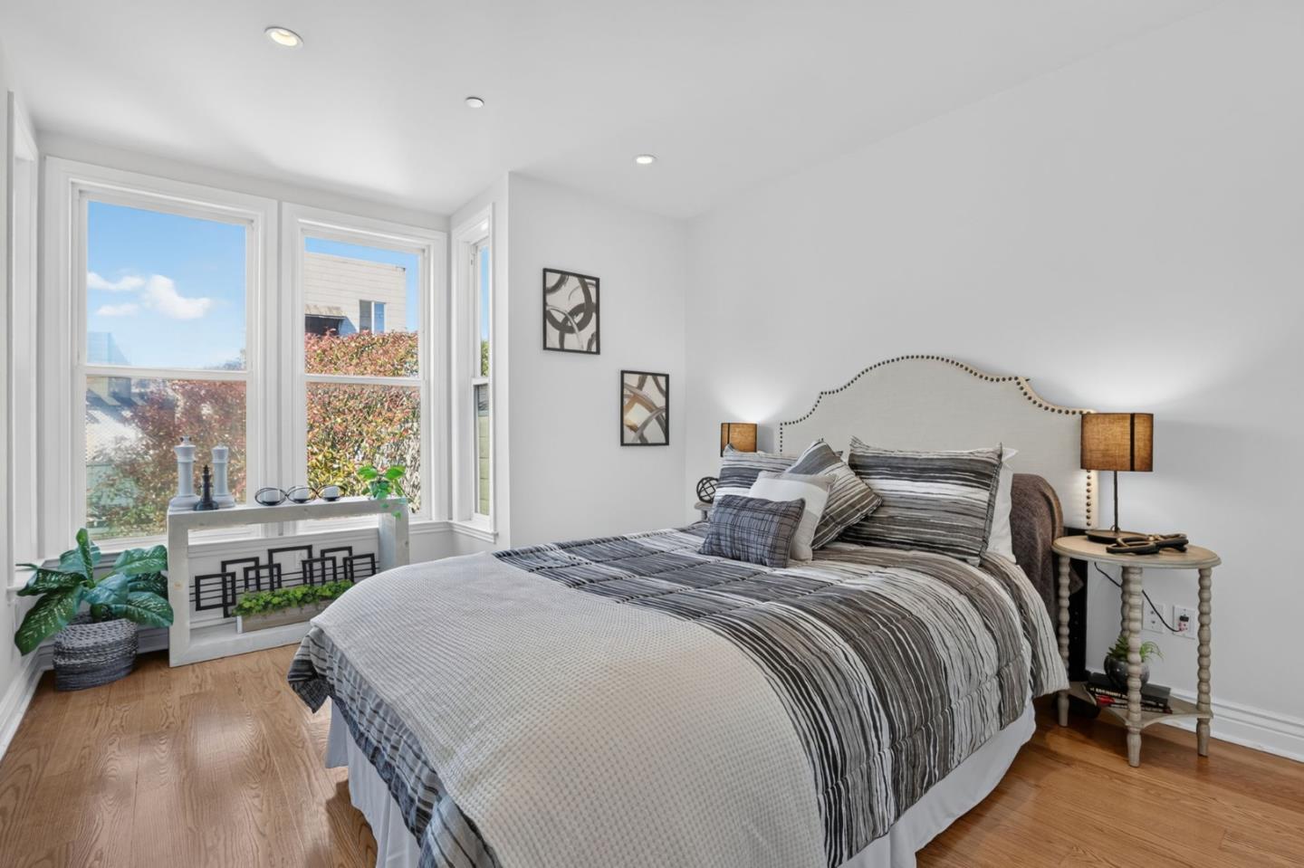 Staged bedroom with a gray-striped bed, upholstered headboard, hardwood floors, and tall sunny windows.