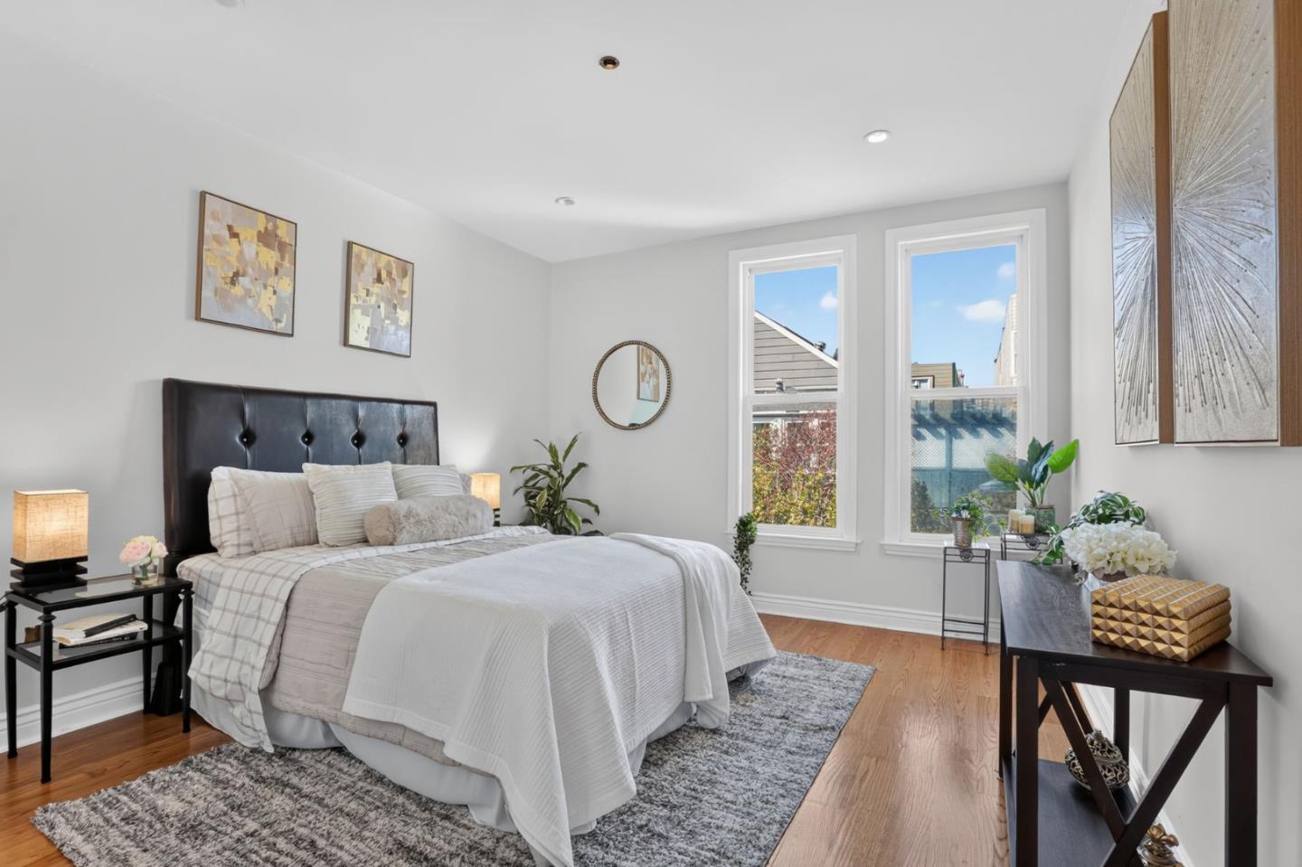 Furnished bedroom with neutral bedding, a black tufted headboard, hardwood floors, and two tall windows.