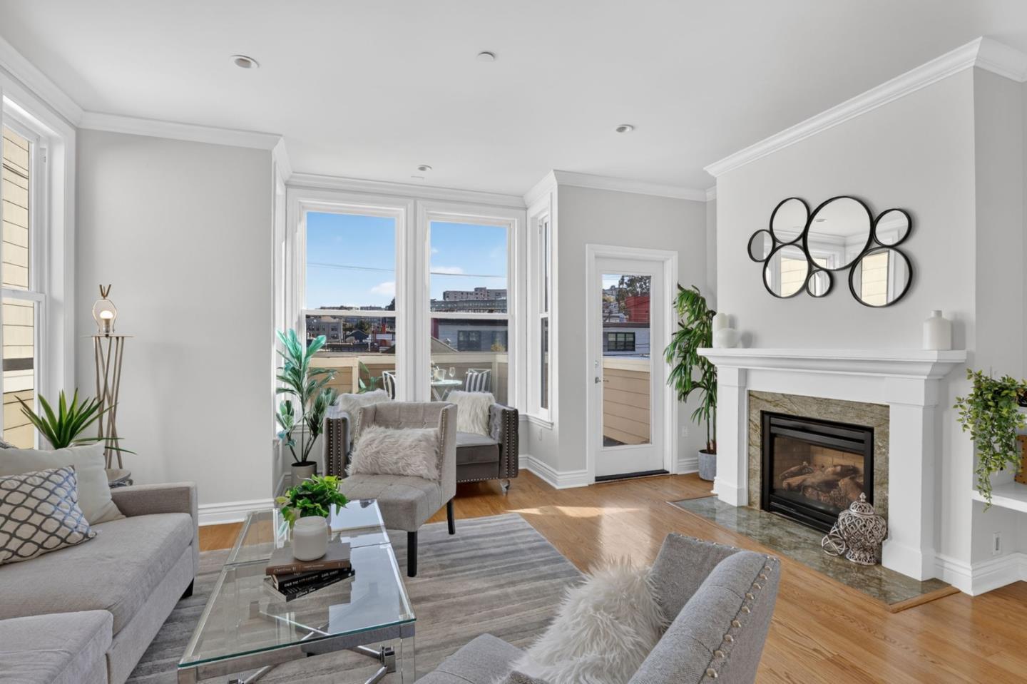Staged living room with gray seating, a glass coffee table, and a white fireplace beside large windows.