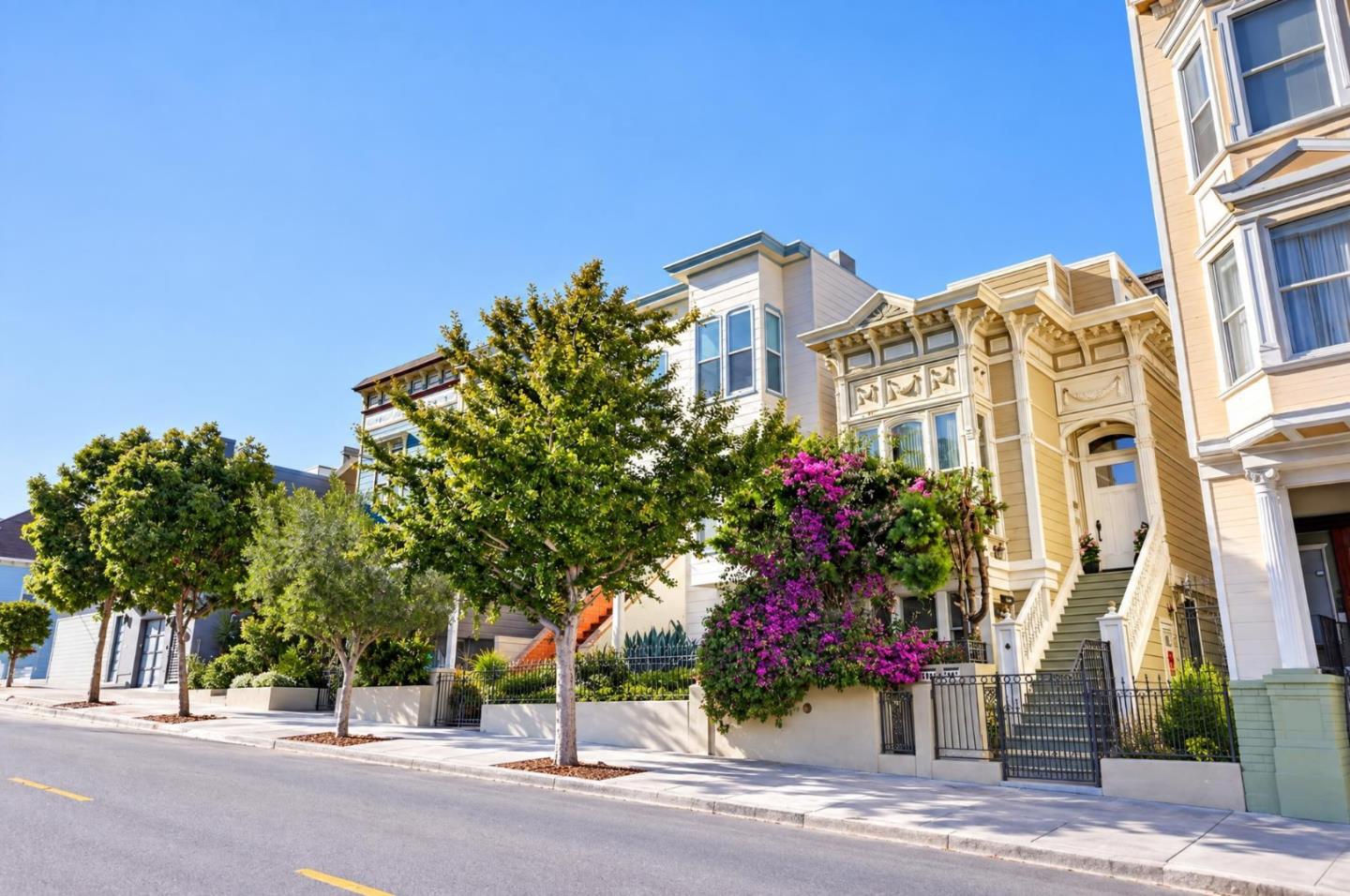 Ornate pale-yellow San Francisco home with front steps, metal fence, and purple flowering shrubs.