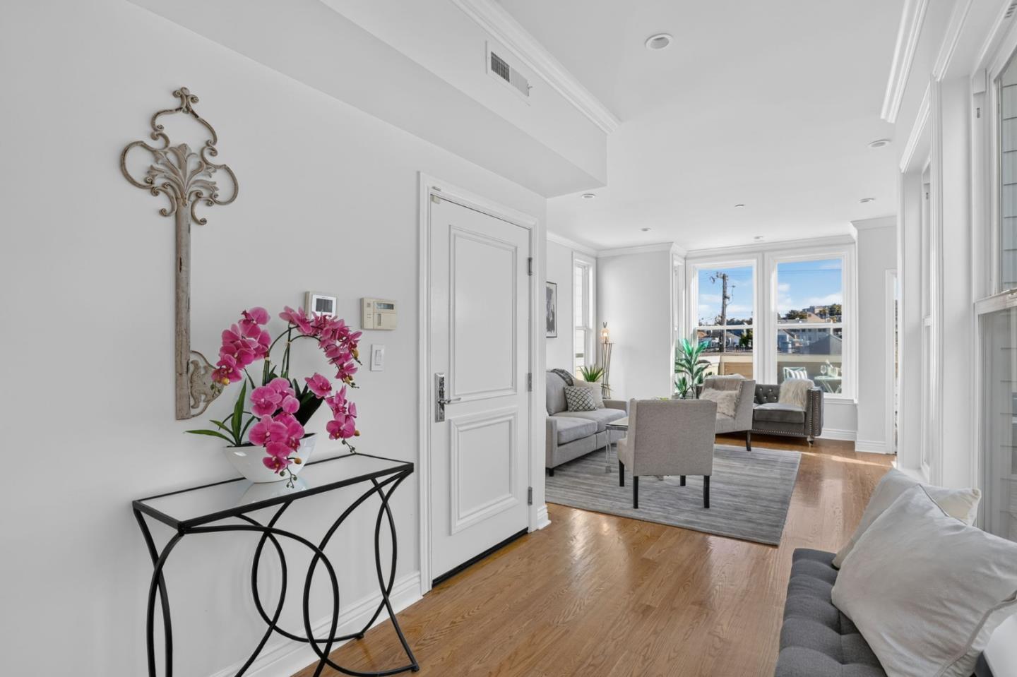 Bright living room with gray seating, hardwood floors, tall windows, and pink orchids by the entry.