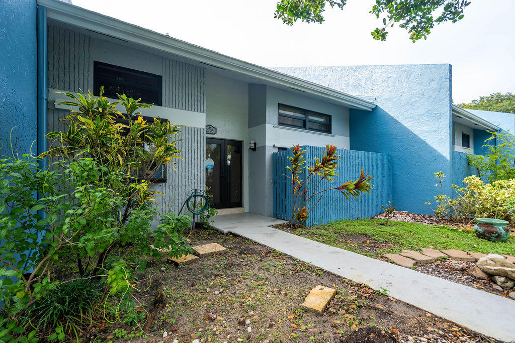 Blue-and-gray residence with a covered entry, blue fence, tropical landscaping, and a walkway to the front door.