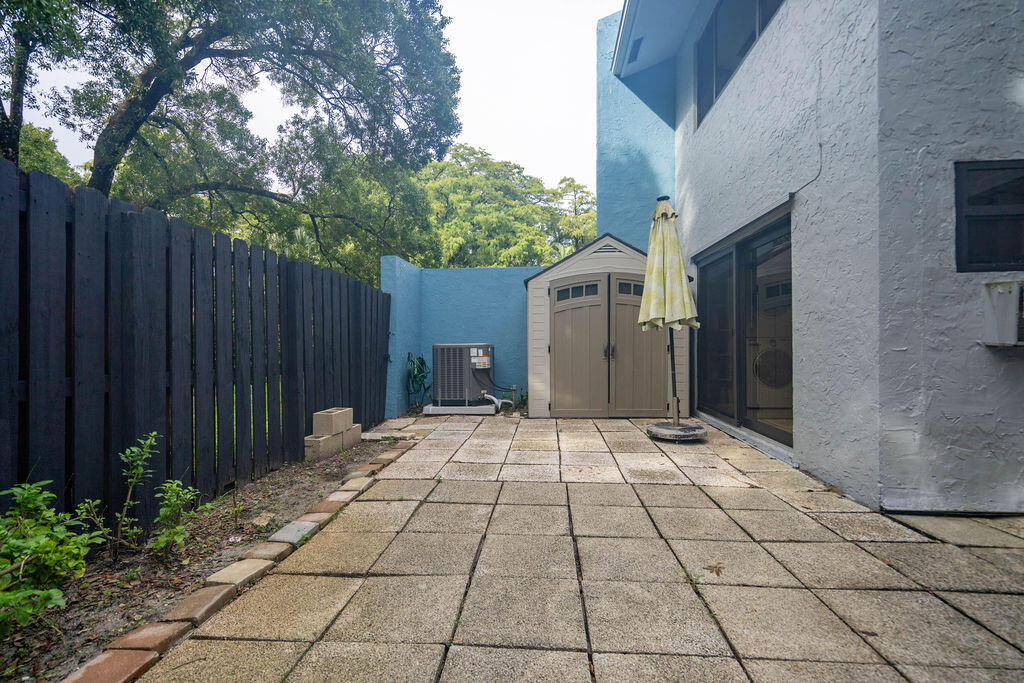 Paved patio beside a white stucco home, enclosed by a tall fence with a storage shed at the back.