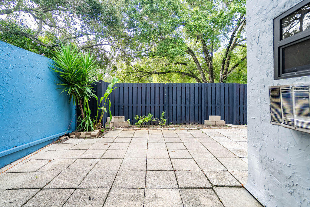 Paved patio with a dark privacy fence, tropical plants, and a window air conditioner.