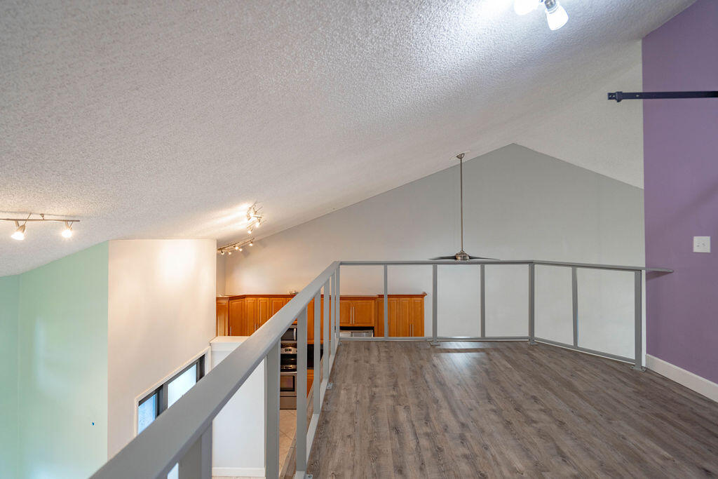 Empty loft with gray wood-look flooring, metal railing, and kitchen with warm wood cabinets below.