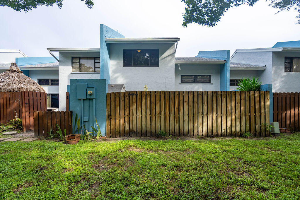 White and blue residential building behind a wooden fence, with a grassy yard and thatched-roof shelter.