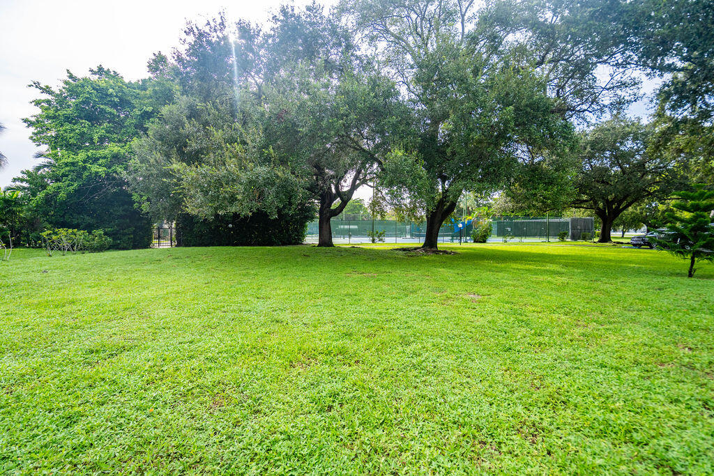 Grassy common area shaded by mature trees, with a fenced tennis court in the background.