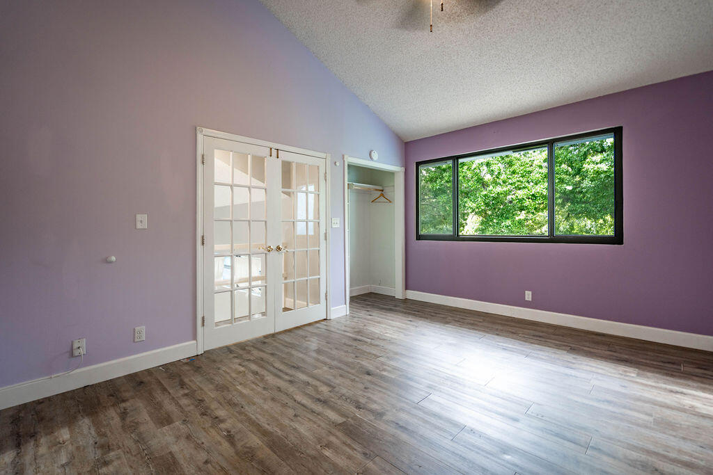 Empty lavender room with wood-look flooring, French doors, and a wide window facing leafy trees.