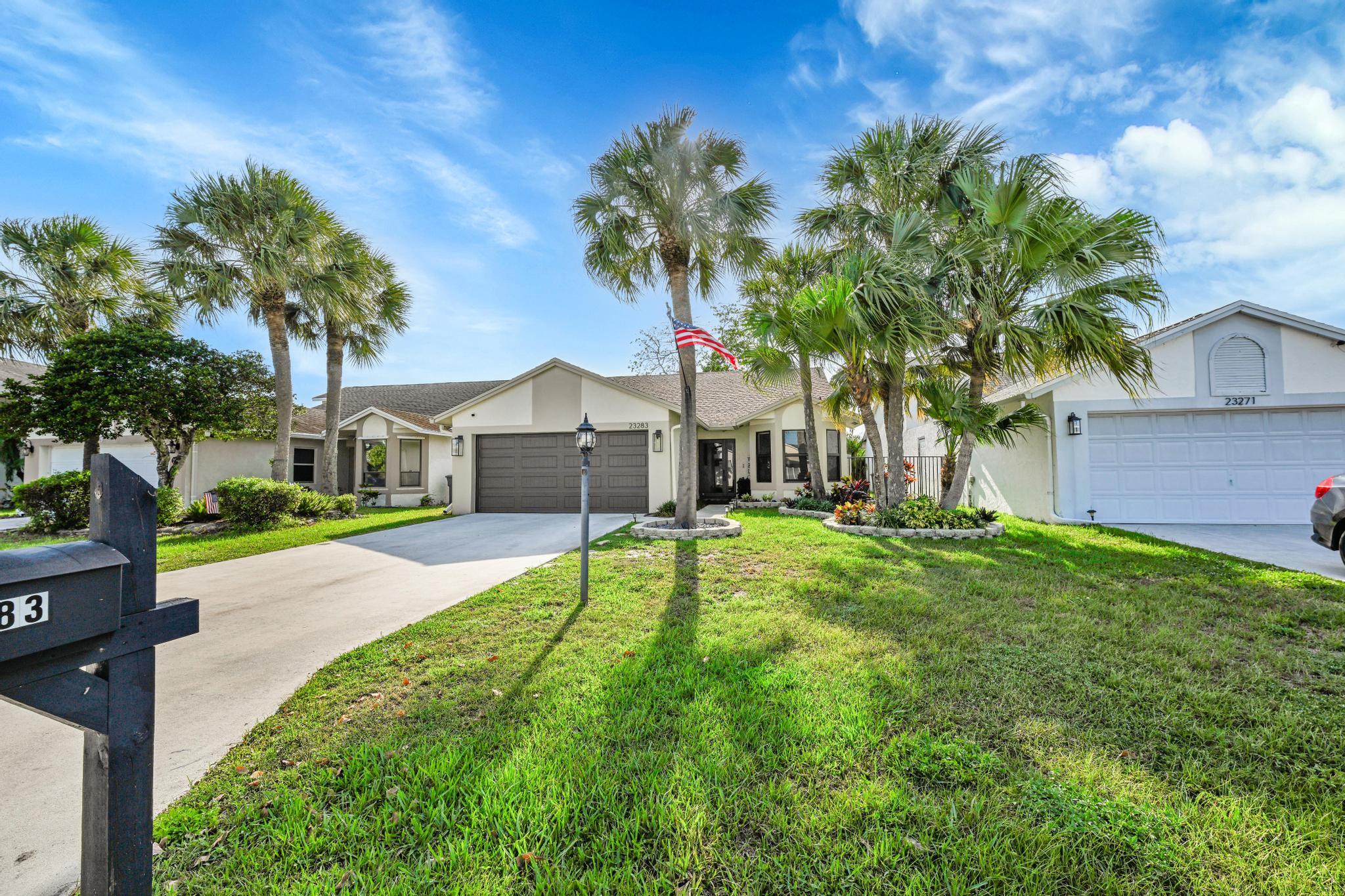 Single-story home with a two-car garage, palm trees, landscaped lawn, and an American flag.
