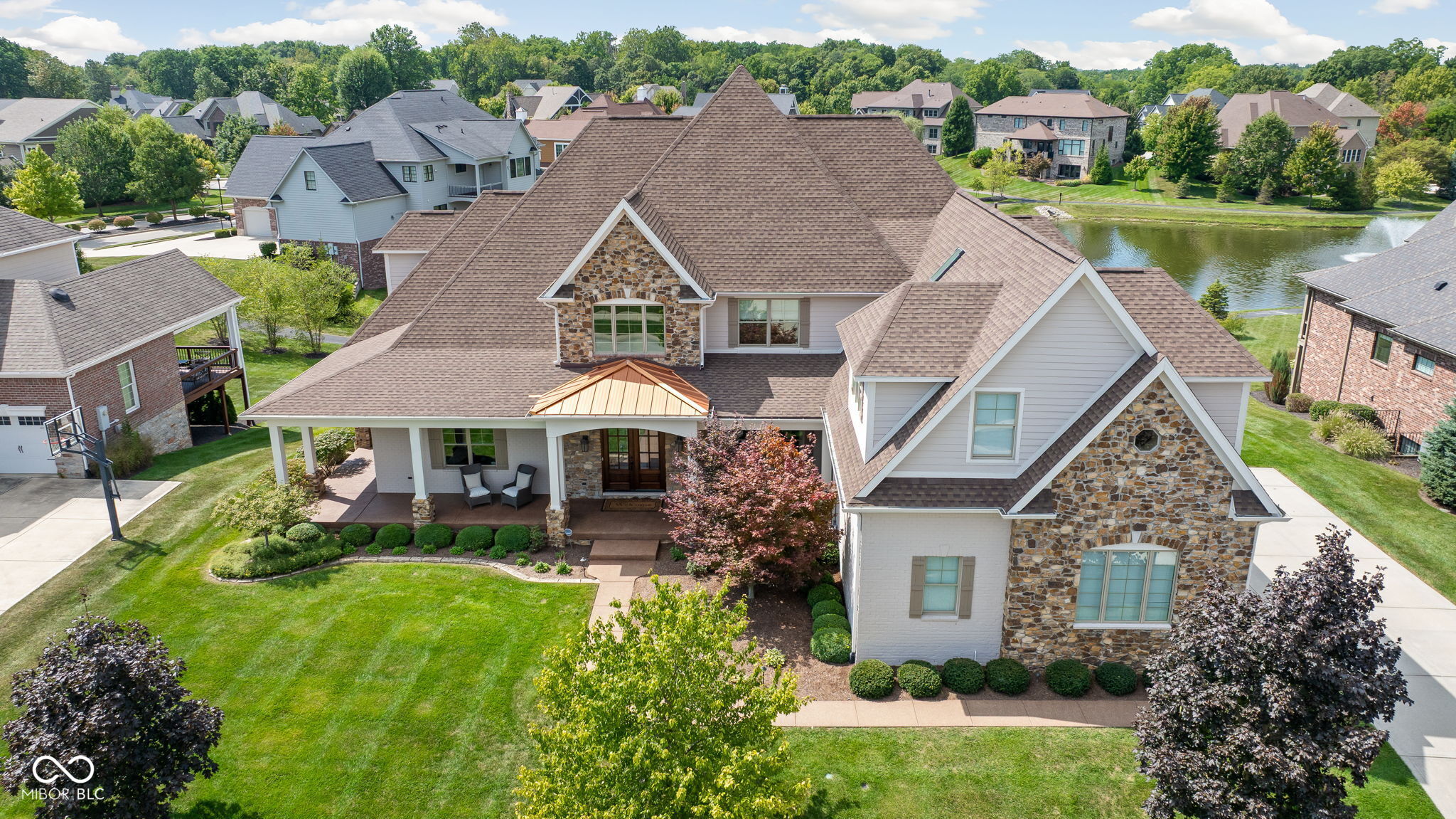 Aerial view of a stone-and-white-sided home beside a neighborhood pond.