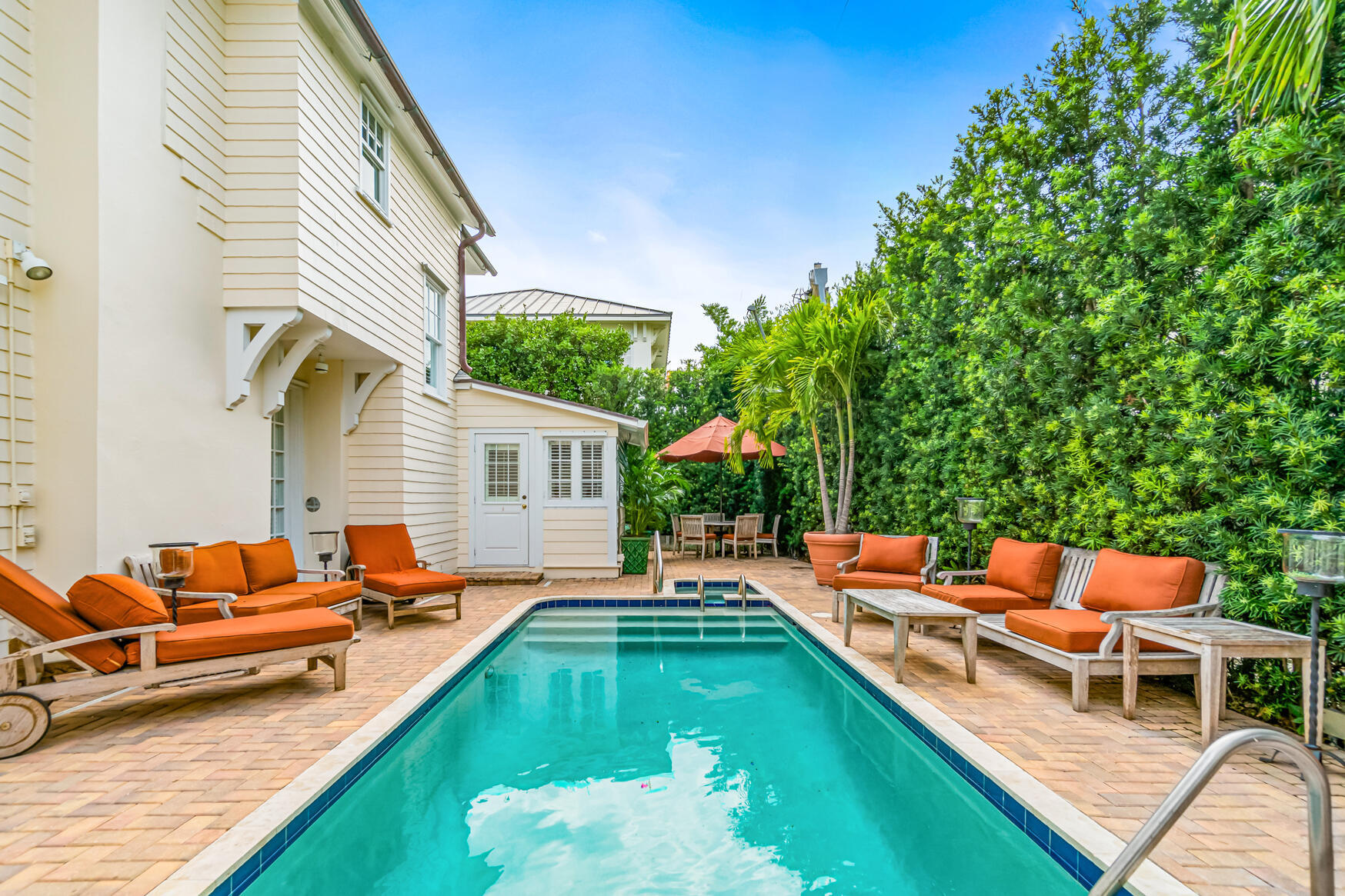 Outdoor pool beside a light-colored house, with orange lounge chairs and tropical landscaping.