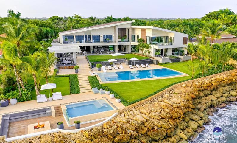 Aerial view of a modern coastal villa with two pools, landscaped lawn, and rocky shoreline.